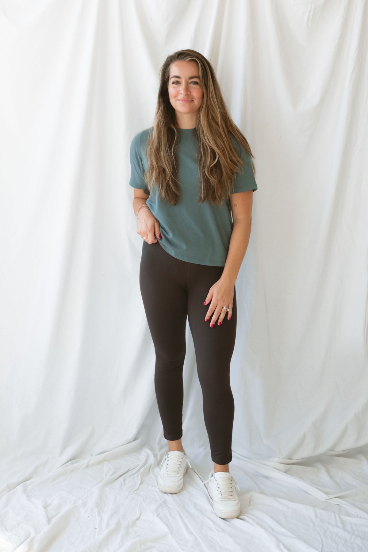 Woman with long light brown air wearing coffee colored leggings with eucalyptus t-shirt and white sneakers on a white background