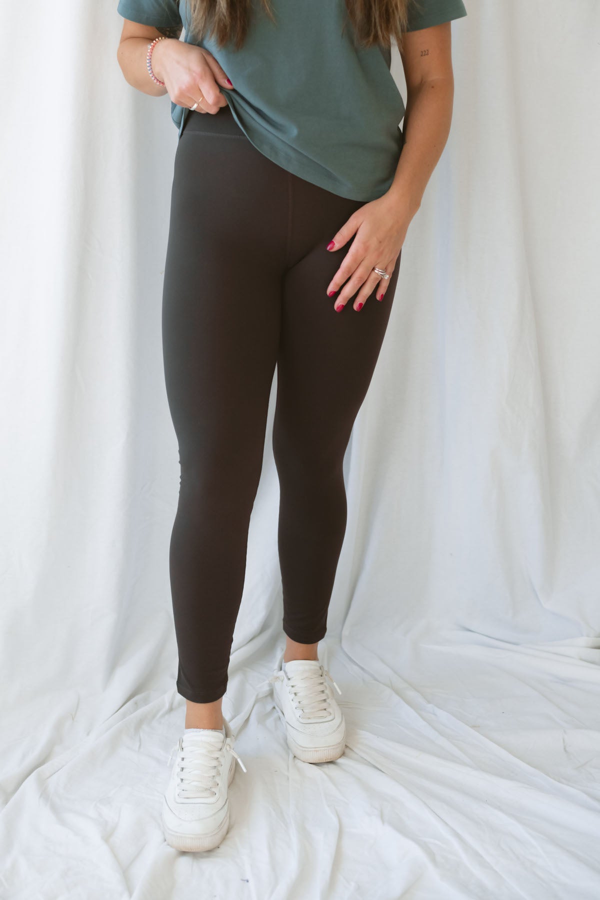 Woman wearing coffee colored leggings with eucalyptus t-shirt and white sneakers on a white background