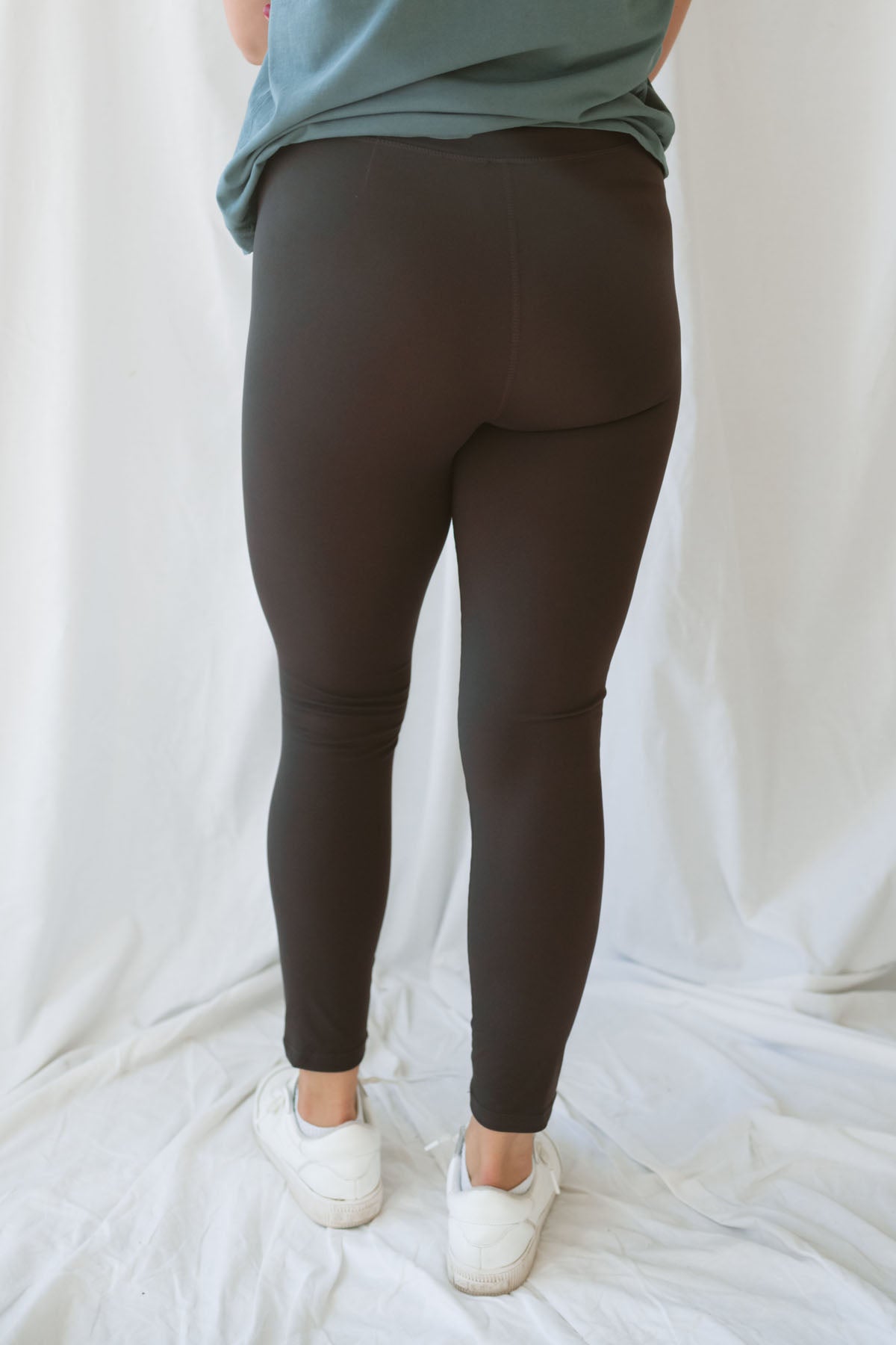 Woman wearing coffee colored leggings with eucalyptus t-shirt and white sneakers on a white background