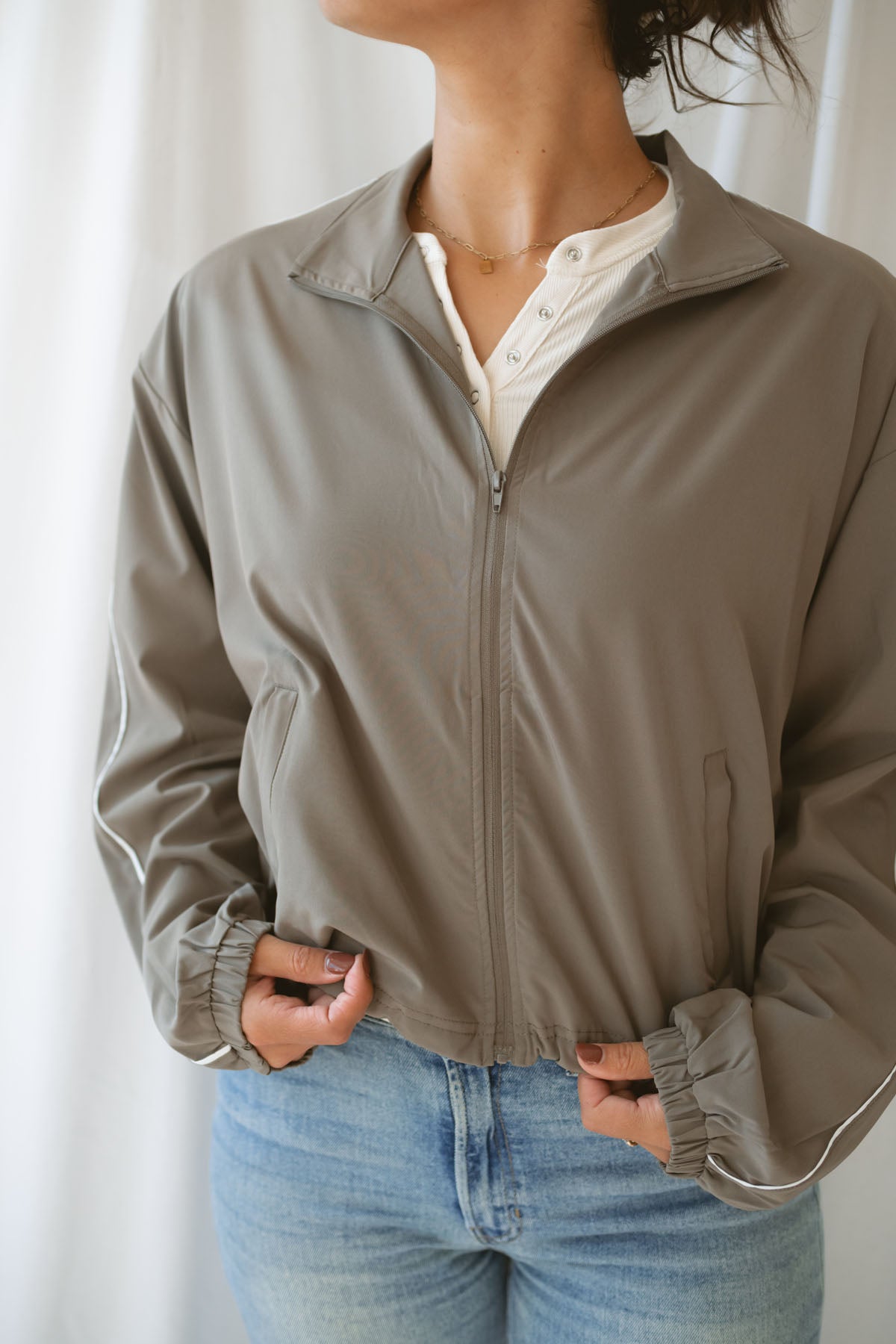 Women with dark hair wearing the truffle active jack with full zip front and pipping detail on the sleeve wearing jeans and standing in front of a white background.