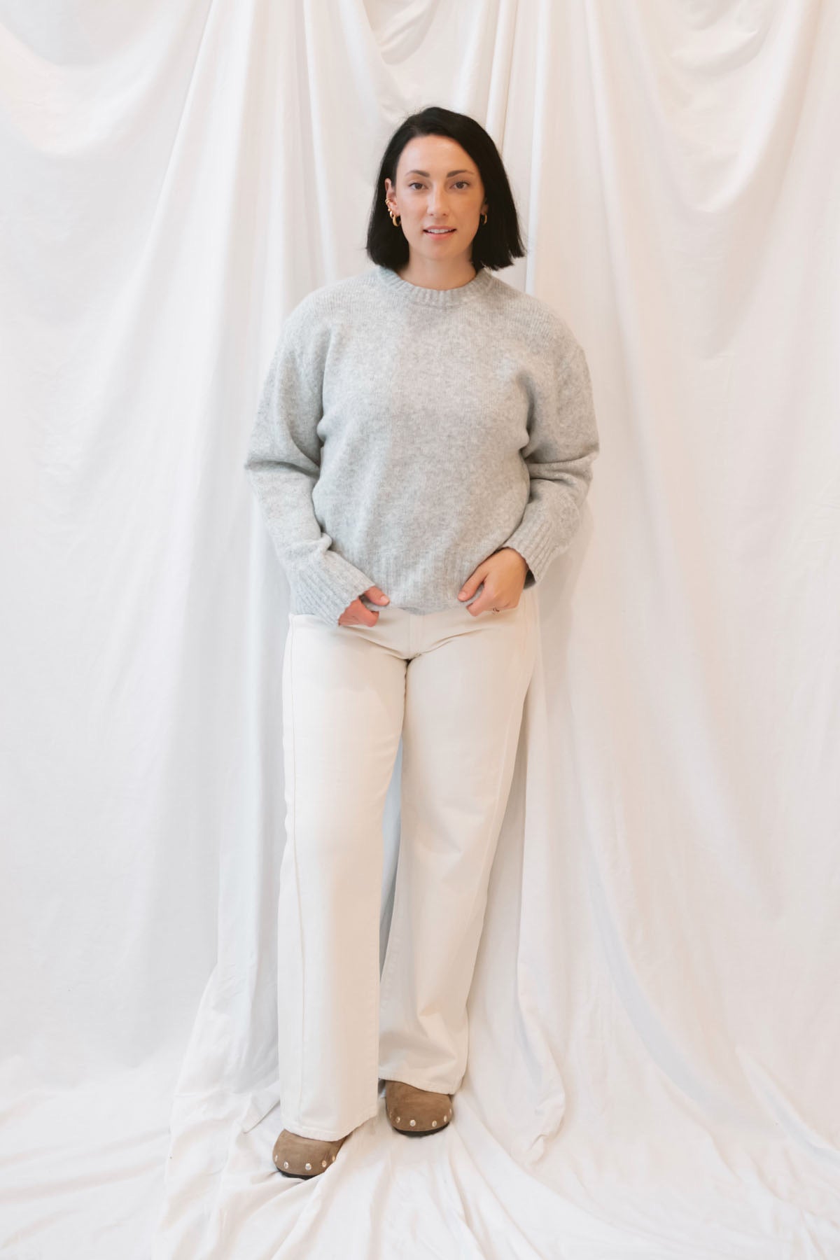 Model is wearing solid heather grey crew neck sweater. Styled with cream twist barrel pants.