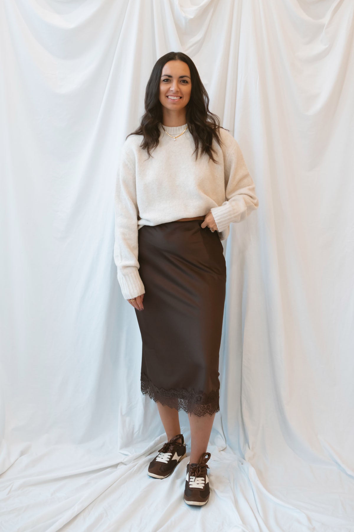 Model wearing a round neck solid sweater in light taupe. Styled with a brown satin skirt with lace trim.