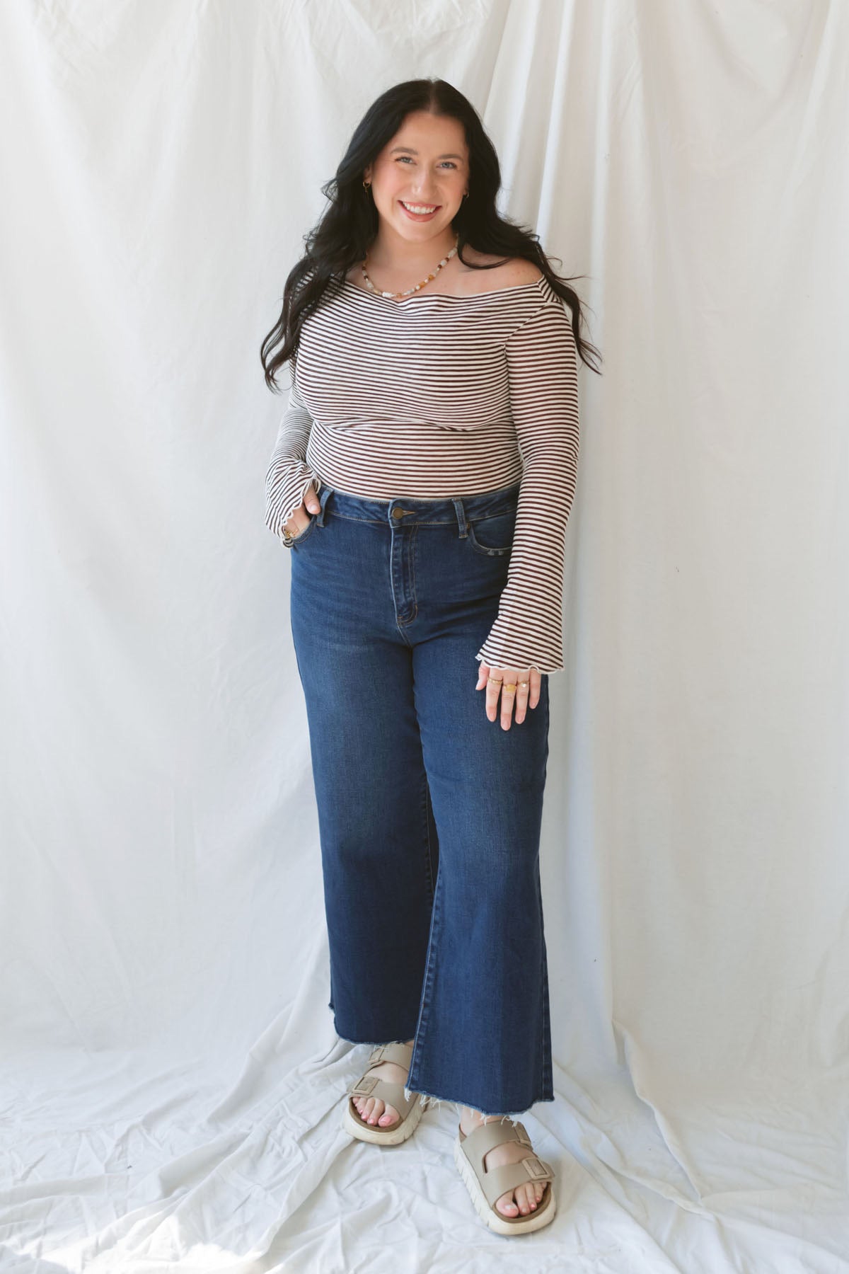 Model wearing the asymmetrical off the shoulder striped top with a pair of basic dark denim and a pop of color in the necklace. Top has lettuce hem detail on the sleeves and a fitted look.