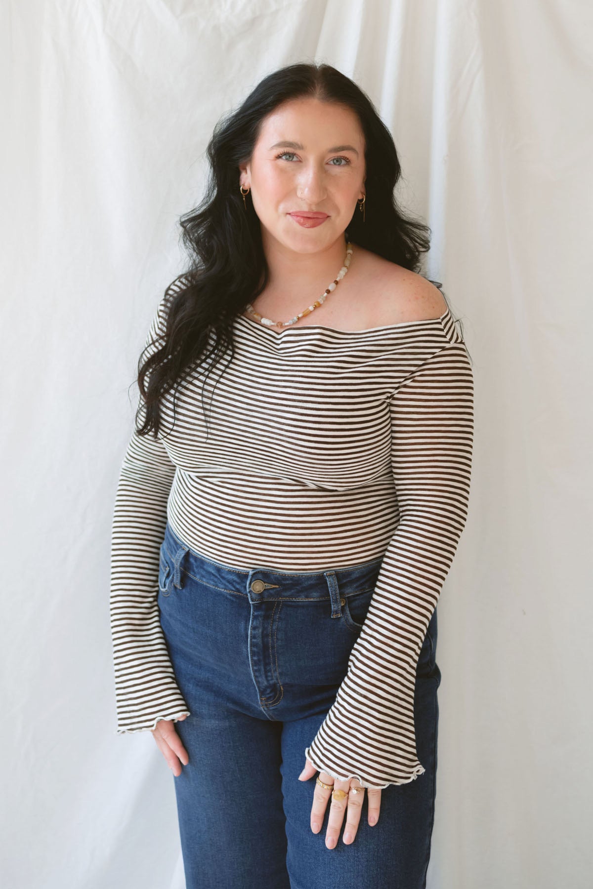 Model wearing the asymmetrical off the shoulder striped top with a pair of basic dark denim and a pop of color in the necklace. Top has lettuce hem detail on the sleeves and a fitted look.