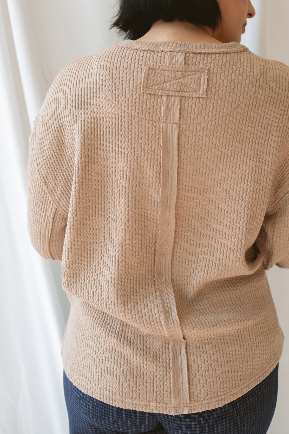 Back view of model wearing textured long sleeve top in color khaki. Details on back include exposed seam.