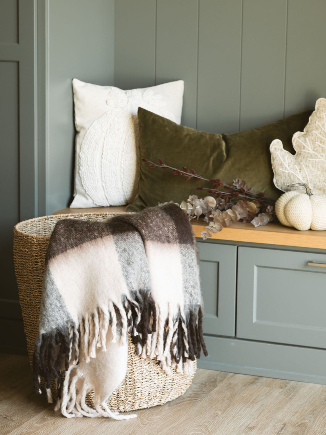 Brown check throw blanket with fringe edges layered into a basket in front of a bench with pillows and soft fabric pumpkins.