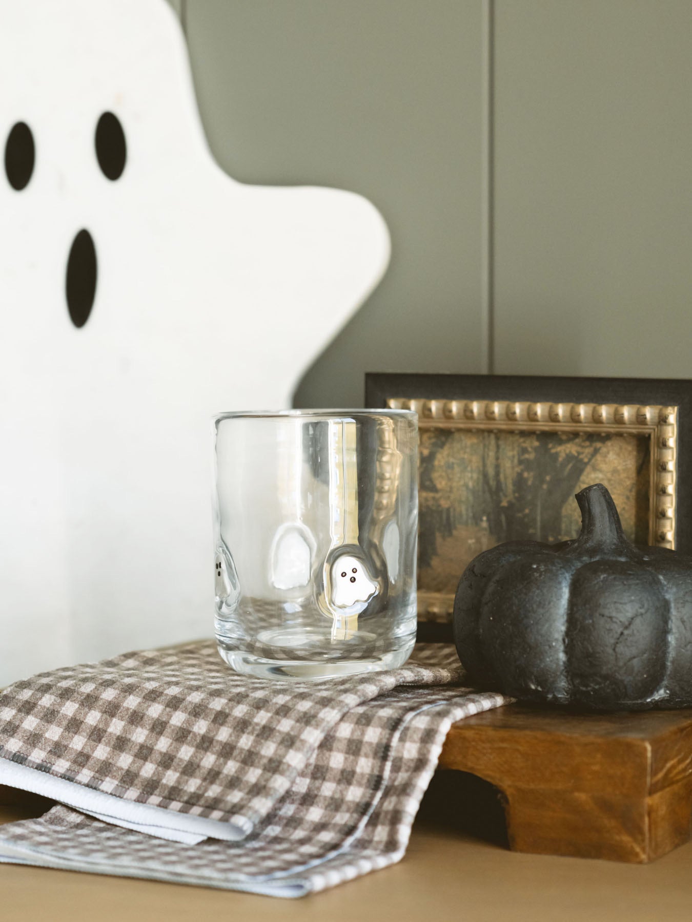 Showing the drinking glass with embedded ghosts sitting on a warm wood board with a checkered geometry towel layered under the glass and a moment created with a marble ghost shaped cheese board leaning behind and a black ceramic pumpkin next to the glass.