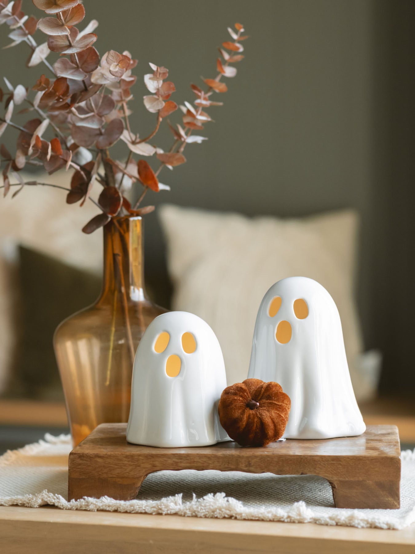 Two sizes of the white ceramic ghosts sitting next to each other on a warm wood riser with a small brown soft pumpkin and the amber glass vase with brown eucalyptus.