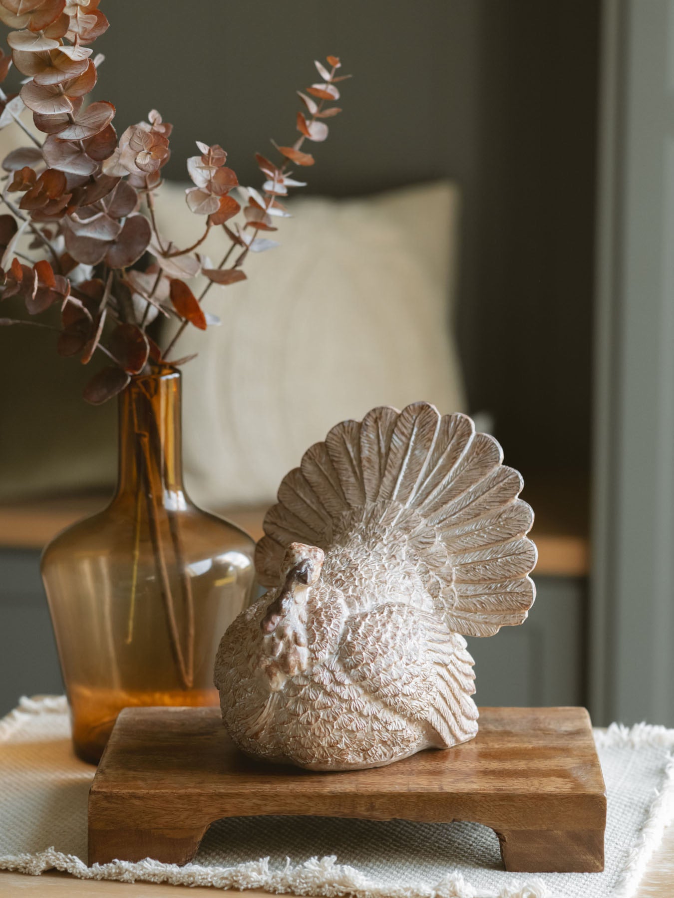 Resin tabletop turkey sitting on a warm wood riser paired with the amber glass vase with a brown eucalyptus stem.