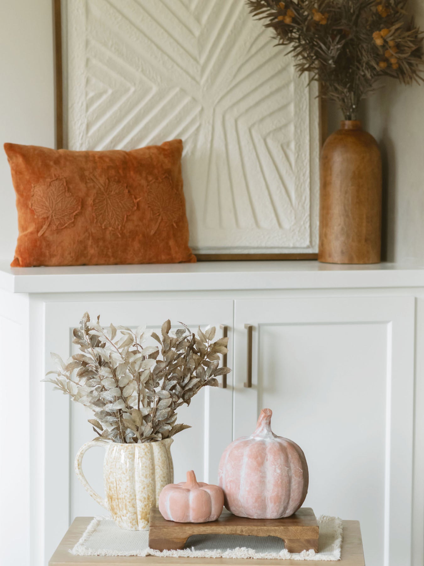 Burnt orange leaf pillow layered on cabinet with large textured art piece and wood vase with sprig.
