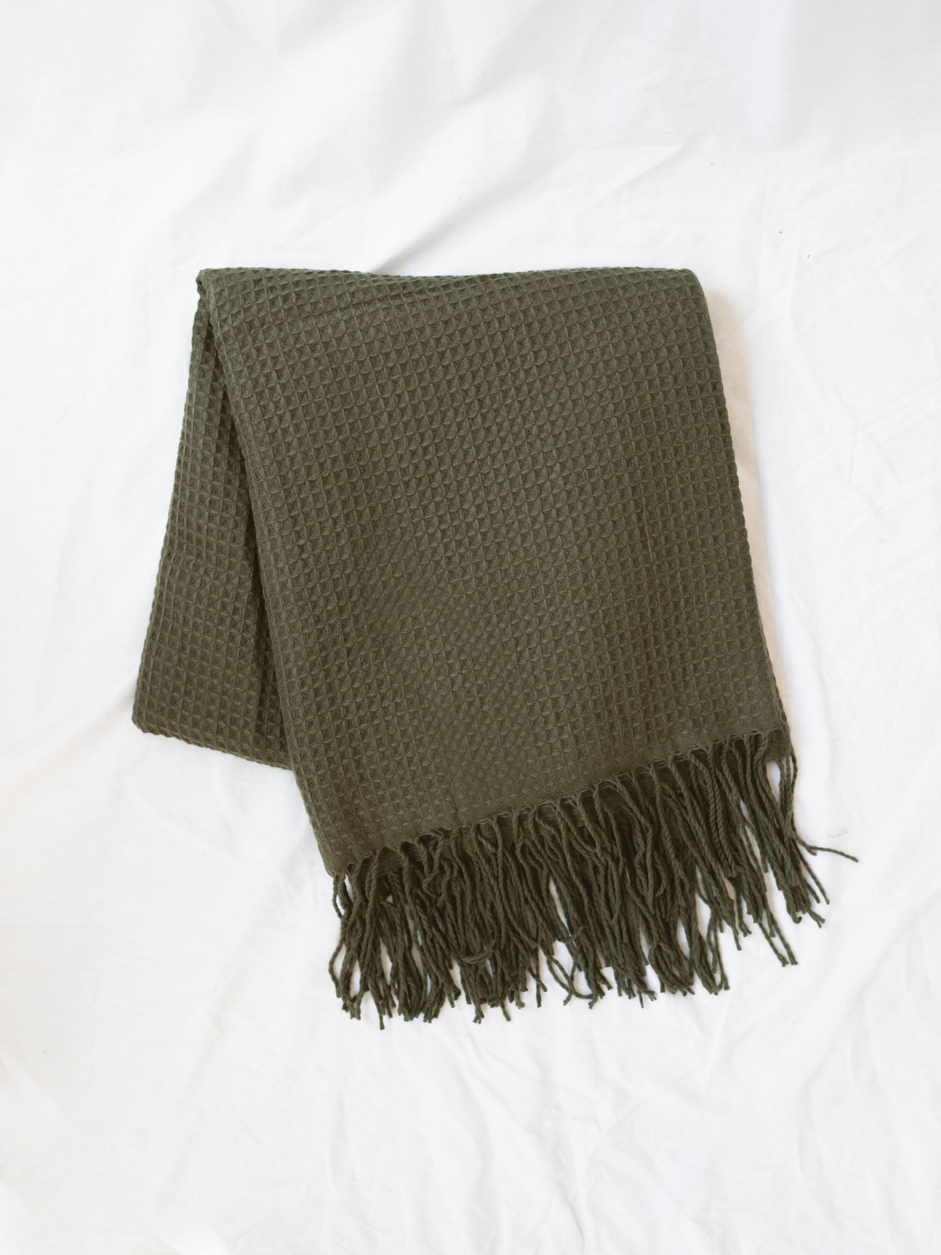 Olive green waffle knit throw blanket folded laying on a white linen backdrop.