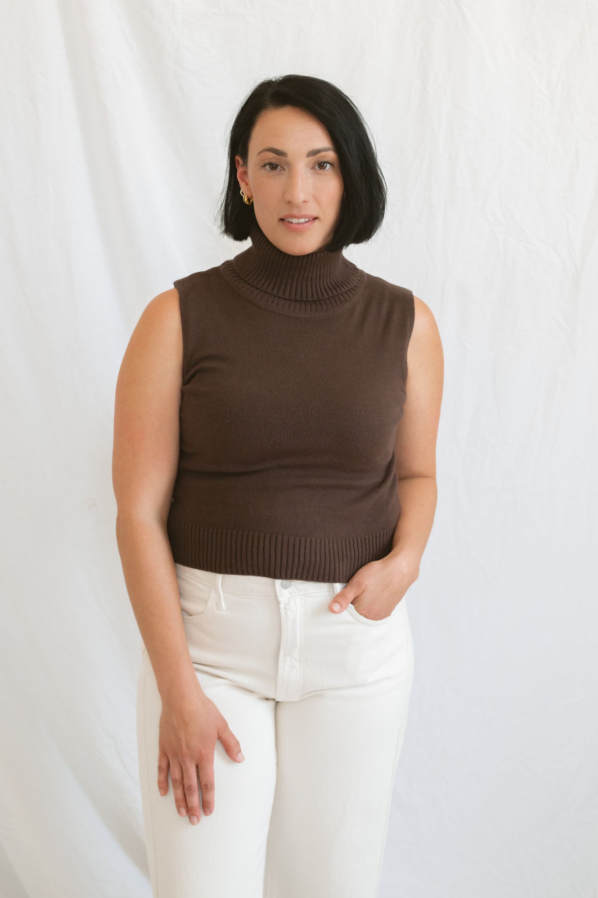 Model wearing the brown sleeveless knit turtleneck. Styled with a cream highwaist denim pant.