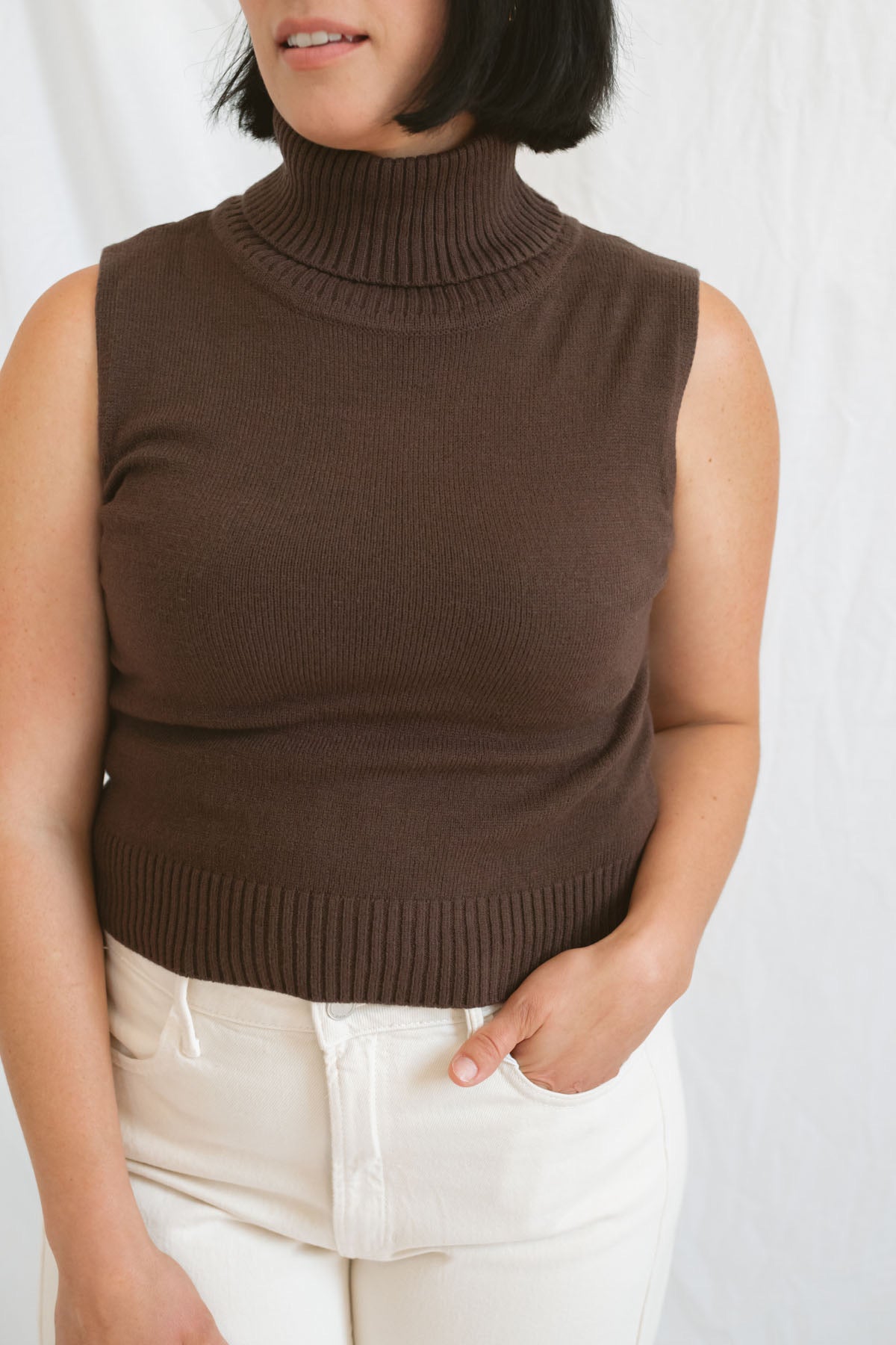 Model wearing the brown sleeveless knit turtleneck. Styled with a cream highwaist denim pant.