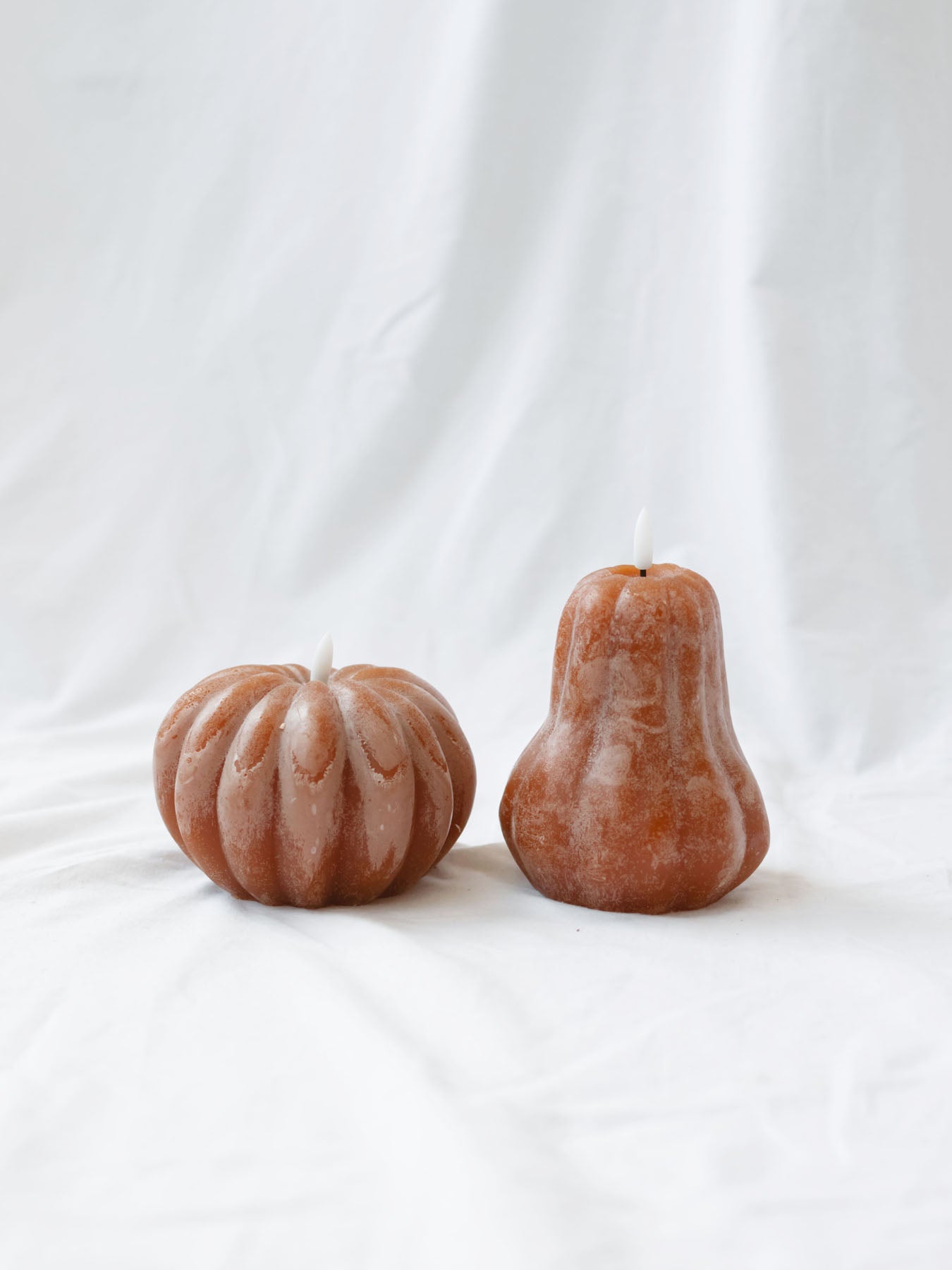 The pumpkin & gourd shape LED candle sitting next to each other on a white linen backdrop.