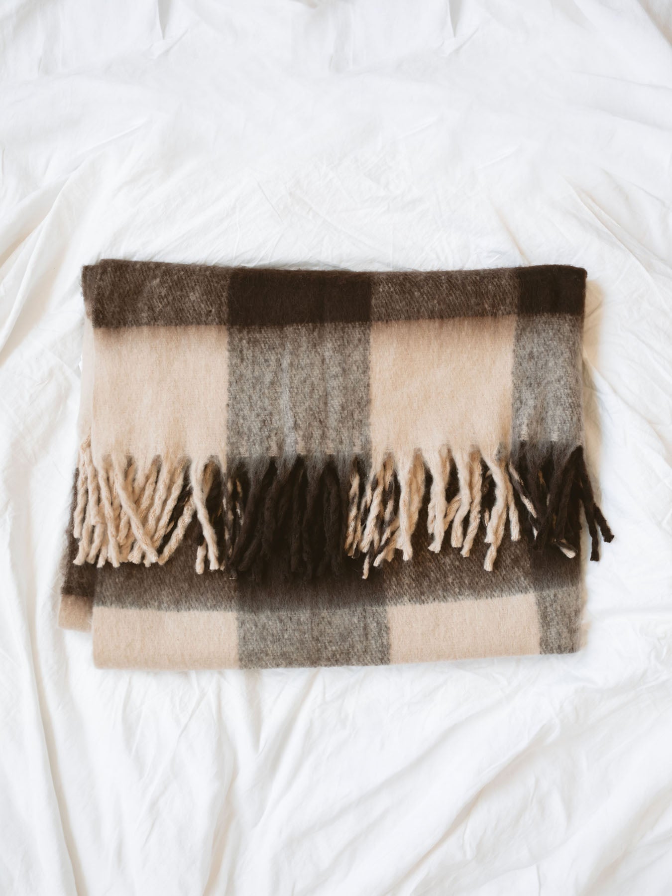 Brown check throw blanket with fringe edges folded laying on white linen backdrop.