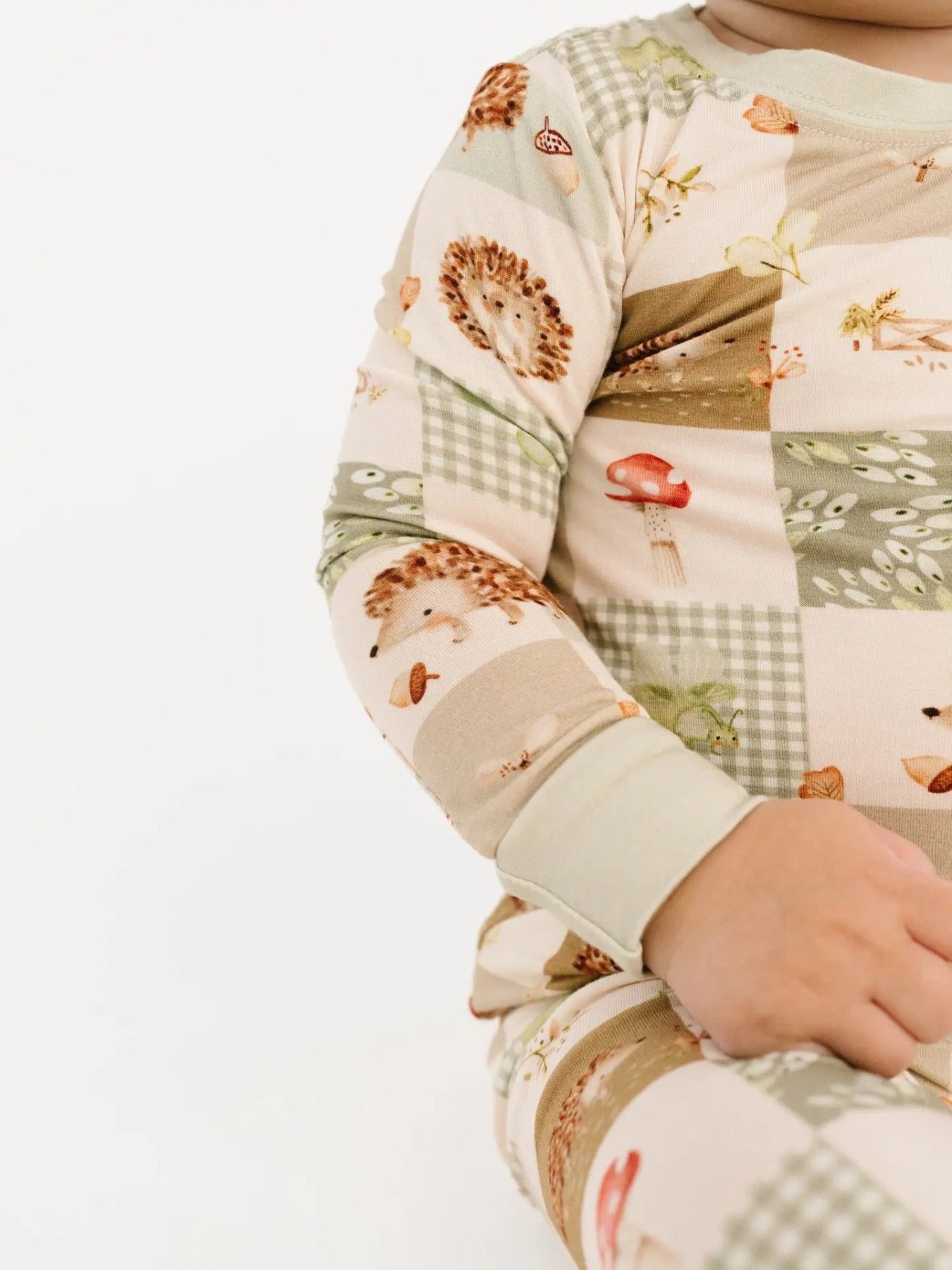 Little boy sitting down wearing a two piece bamboo pajama set with patchwork detail. There are hedgehogs, gingham, mushrooms, acorns, and leaf details all over. 