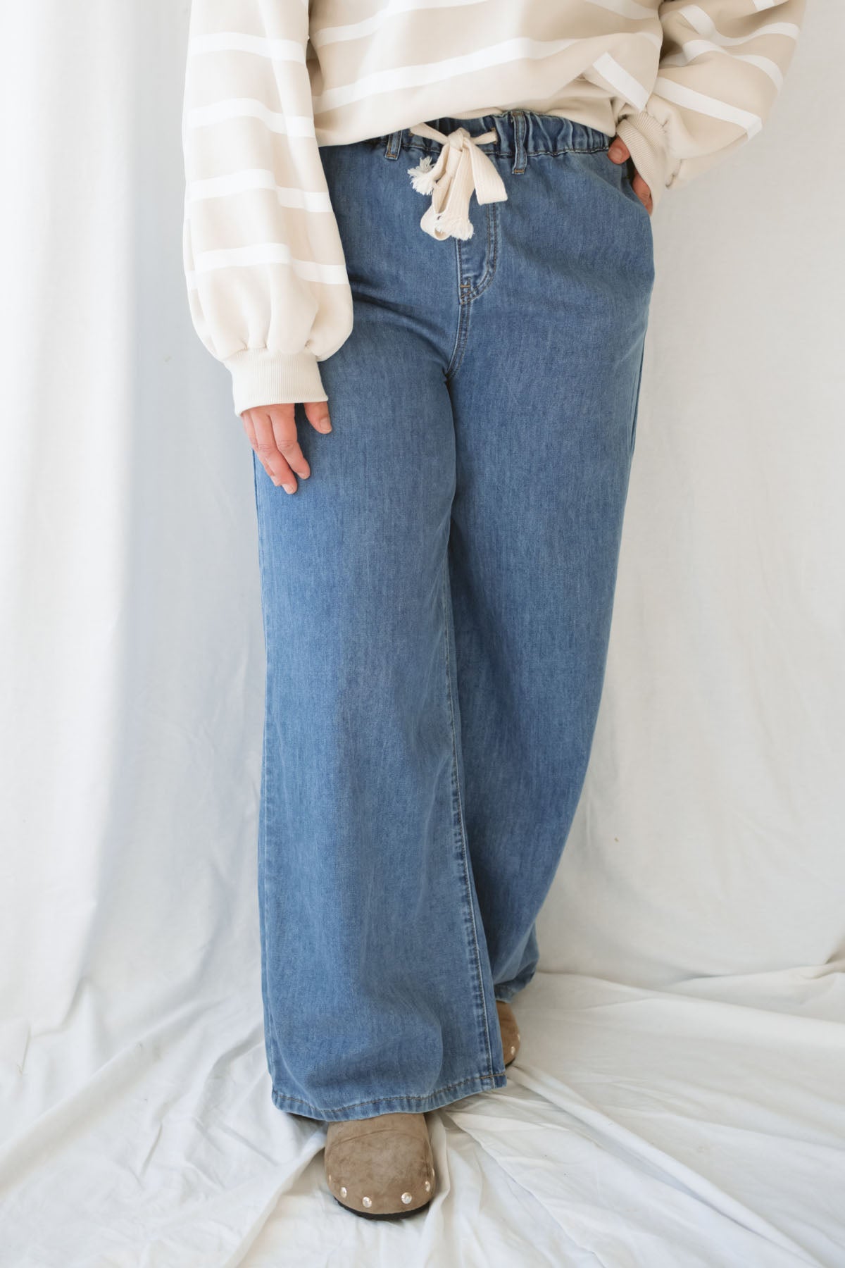Woman wearing wide leg medium wash denim with drawstring waistband with taupe sweatshirt on a white background.
