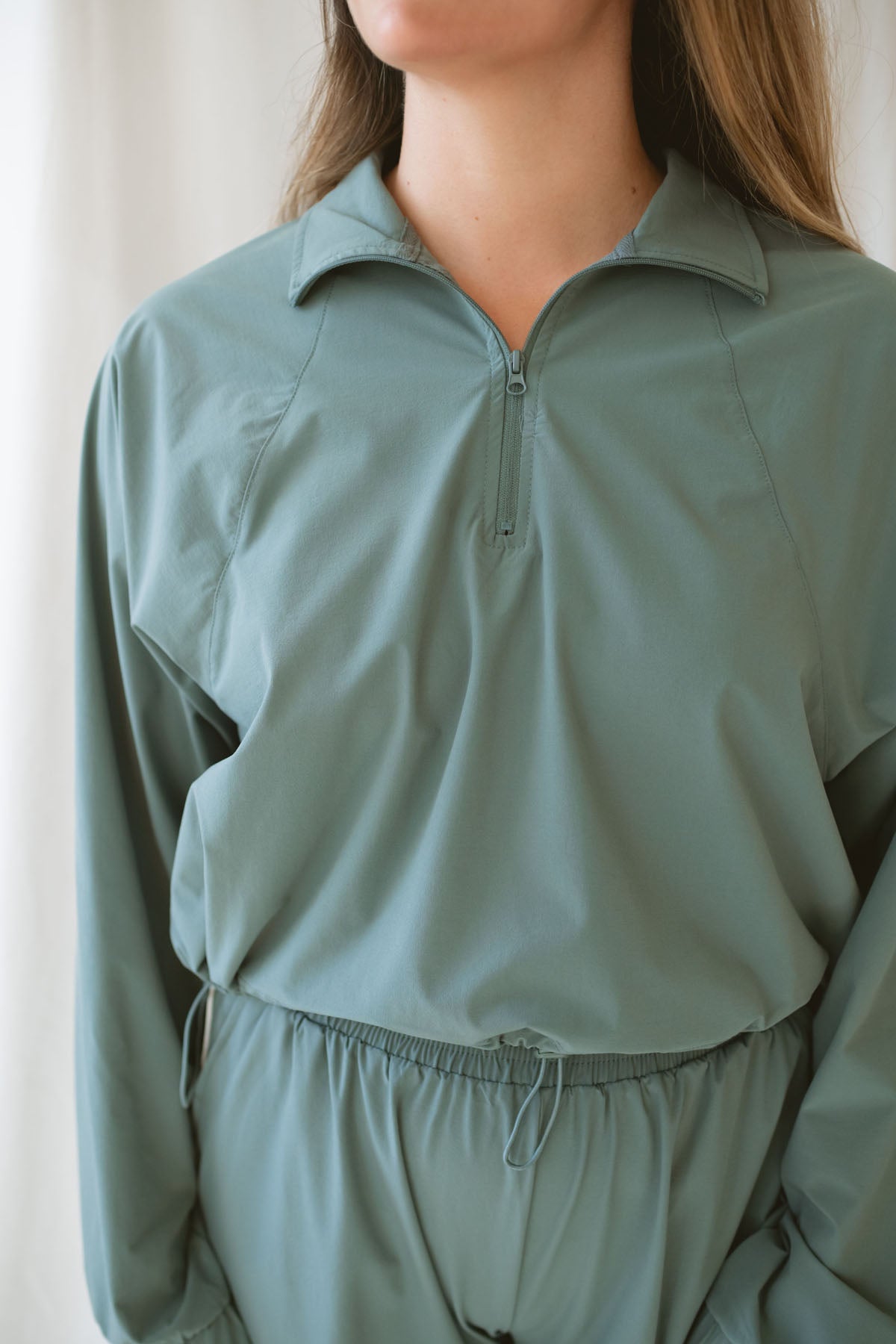 Woman with light brown hair wearing a eucalyptus half zip active jacket with collar wearing matching pants on a white background 