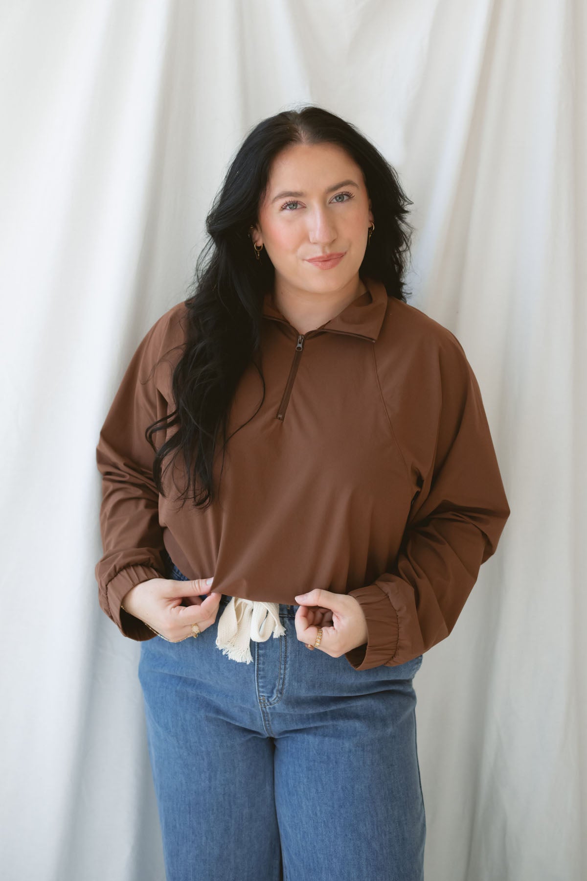 Woman wearing the half zip active jacket in cocoa with medium wash denim and standing in front of a white background 