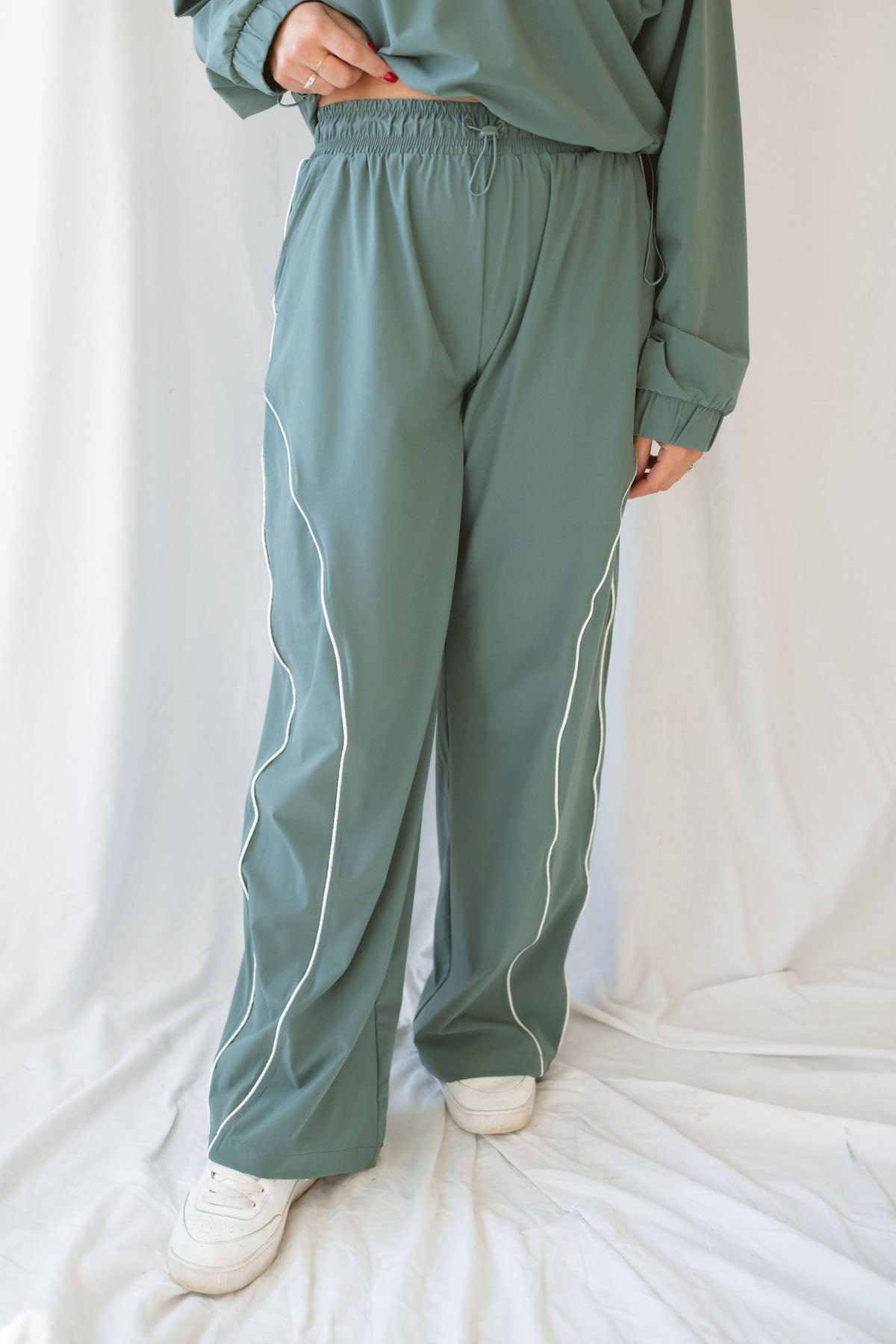 Woman wearing eucalyptus green wide-leg track pants with white contrast piping and an elastic drawstring waistband.