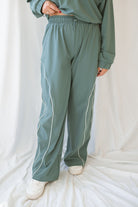 Woman wearing eucalyptus green wide-leg track pants with white contrast piping and an elastic drawstring waistband.