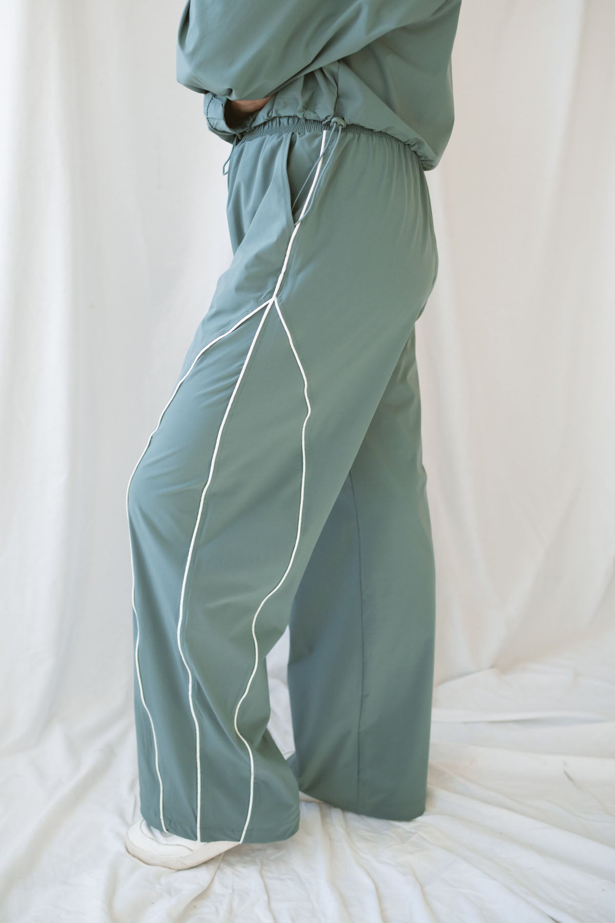 Woman wearing eucalyptus green wide-leg track pants with white contrast piping and an elastic drawstring waistband.