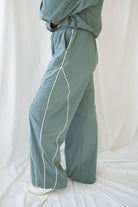 Woman wearing eucalyptus green wide-leg track pants with white contrast piping and an elastic drawstring waistband.