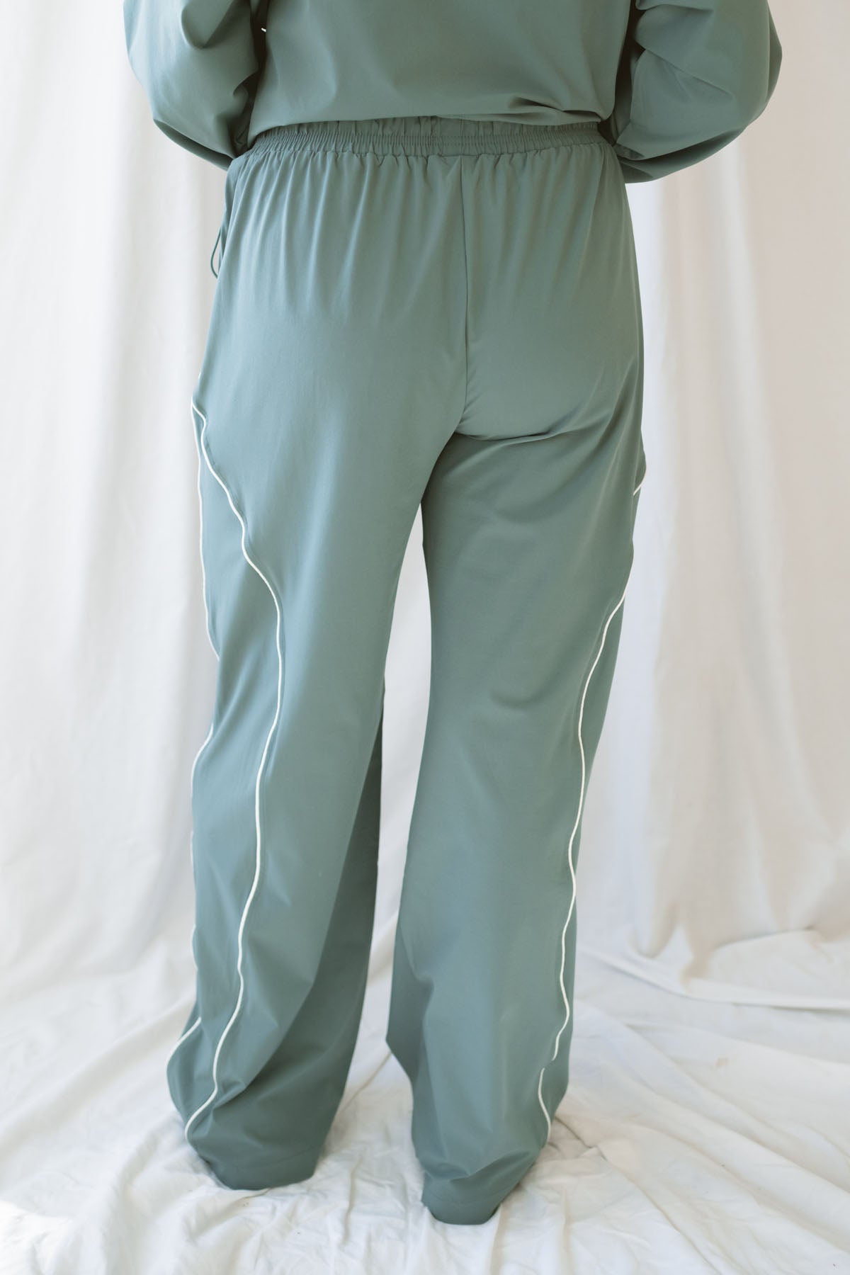 Woman wearing eucalyptus green wide-leg track pants with white contrast piping and an elastic drawstring waistband.