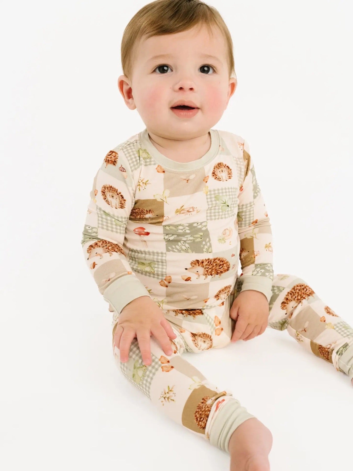 Little boy sitting down wearing a two piece bamboo pajama set with patchwork detail. There are hedgehogs, gingham, mushrooms, acorns, and leaf details all over. 