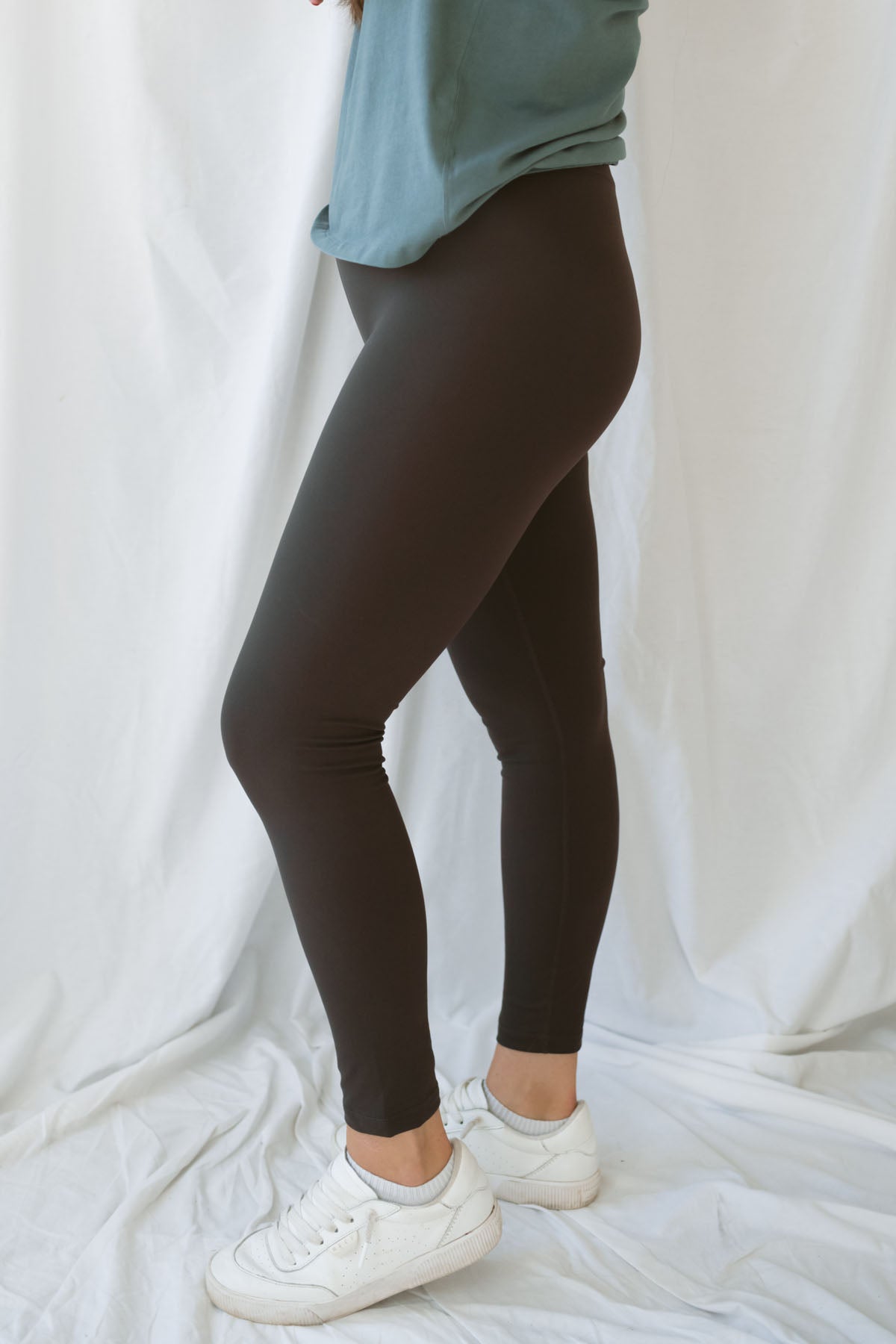 Side Photo of Woman wearing coffee colored leggings with eucalyptus t-shirt and white sneakers on a white background 