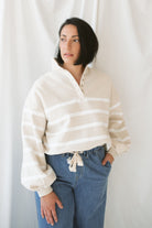 woman with dark hair wearing a taupe button up sweatshirt with white stripe detail wearing jeans on a white background.