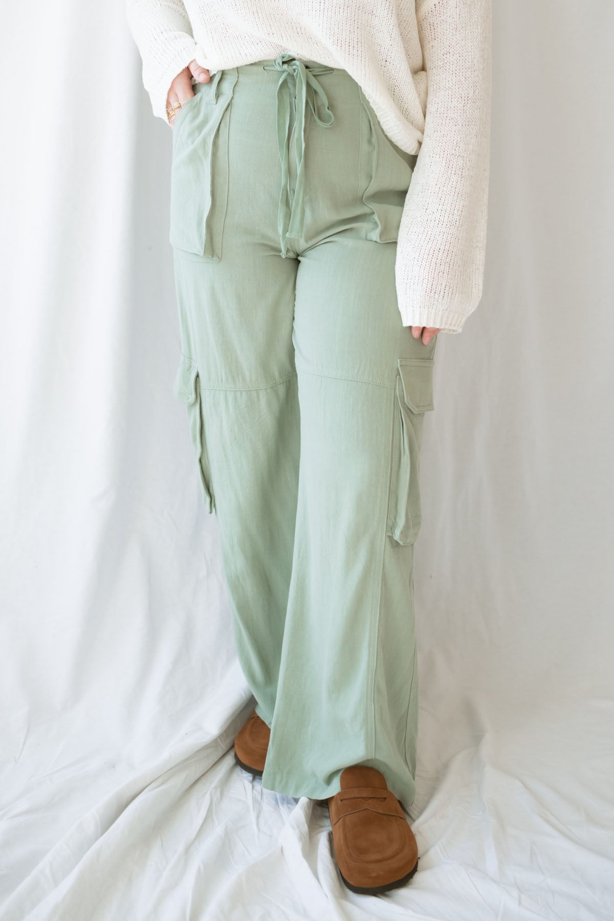 Woman wearing sage green relaxed wide-leg utility pants with oversized cargo pockets and an adjustable drawstring waist.