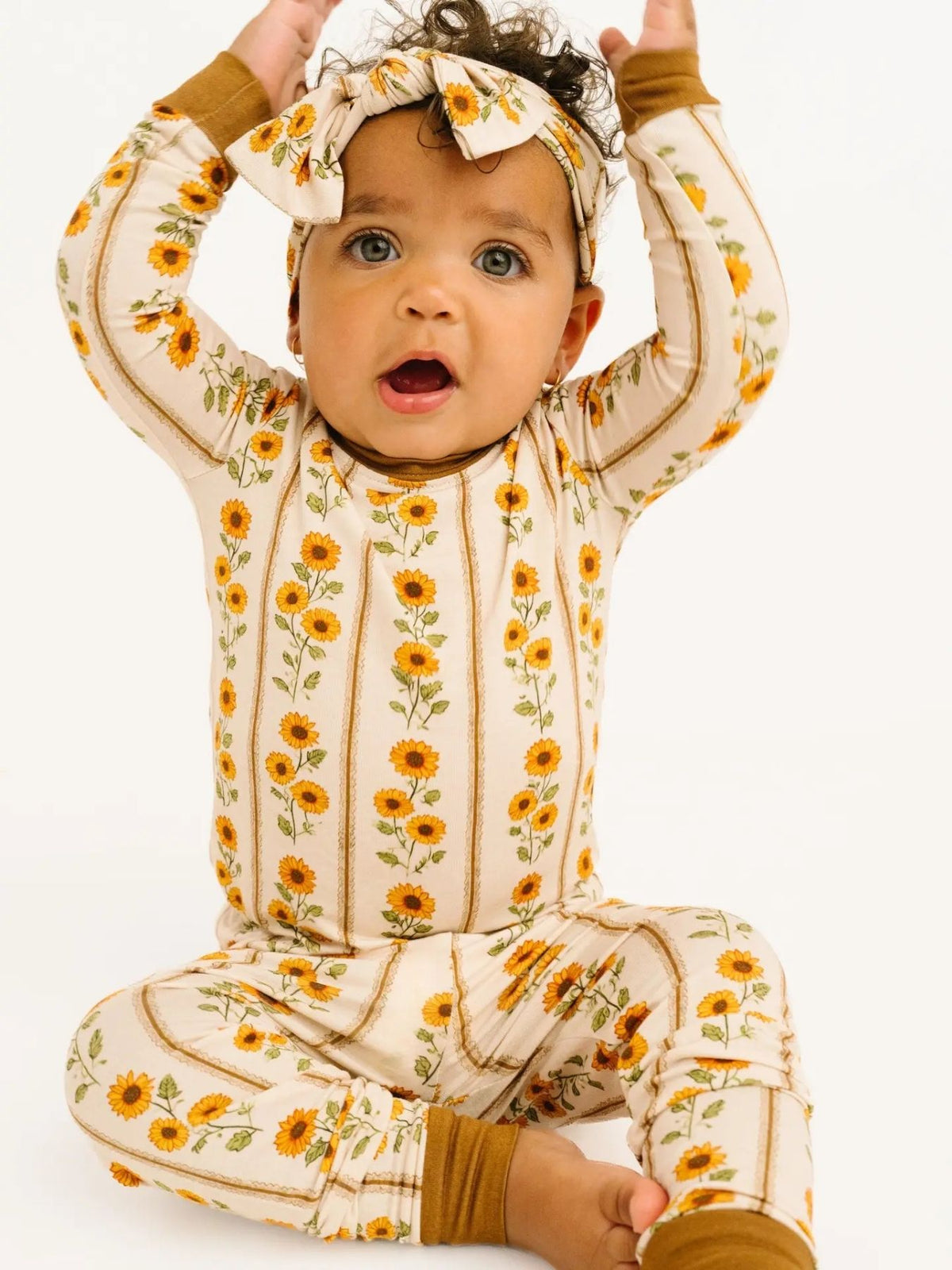 Toddler wearing two-piece pajama set features rows of golden sunflowers on a soft cream background, finished with rich mustard cuffs. Sitting in front of a white background.