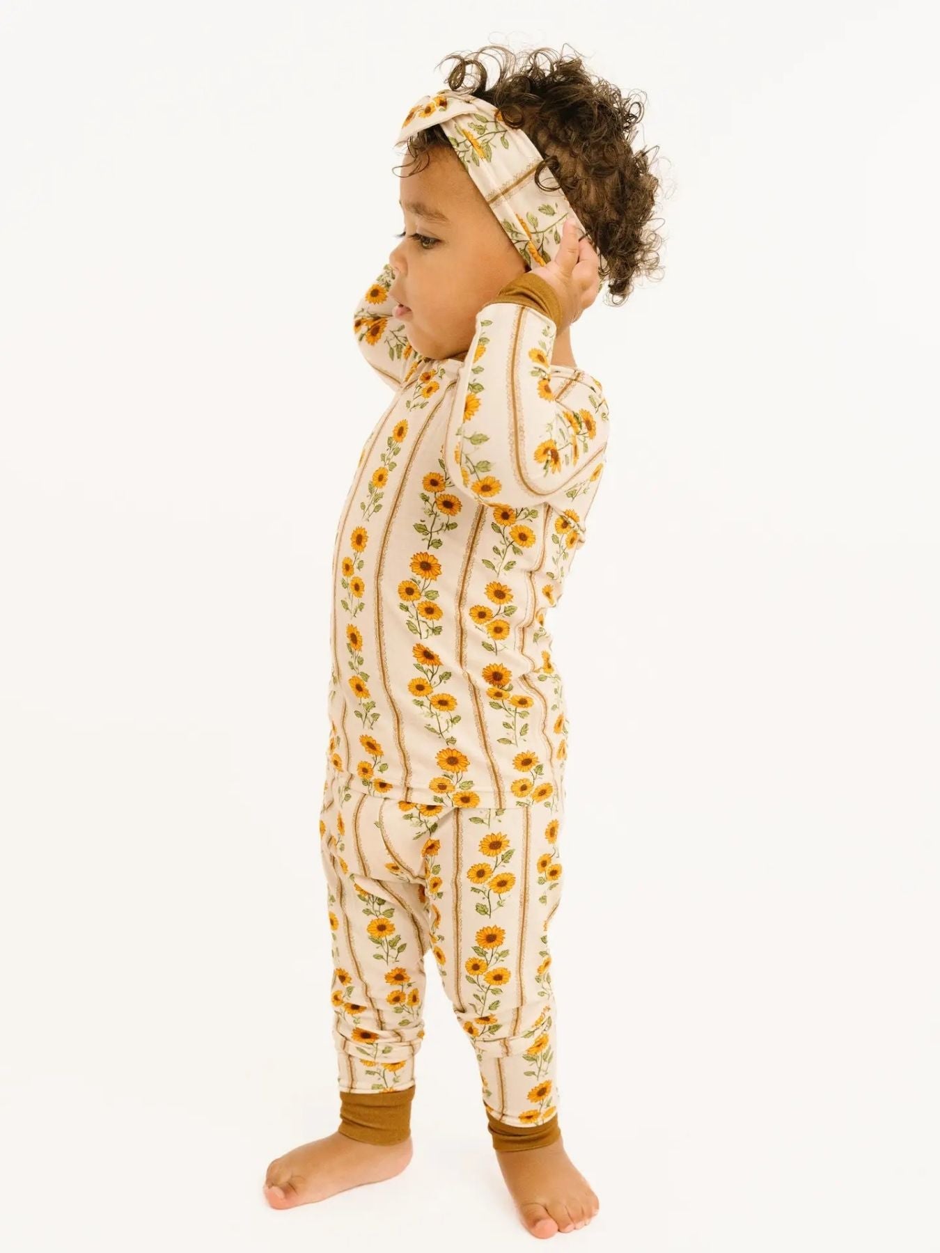 Toddler wearing a two-piece pajama set features rows of golden sunflowers on a soft cream background, finished with rich mustard cuffs. Standing in front of a white background. 