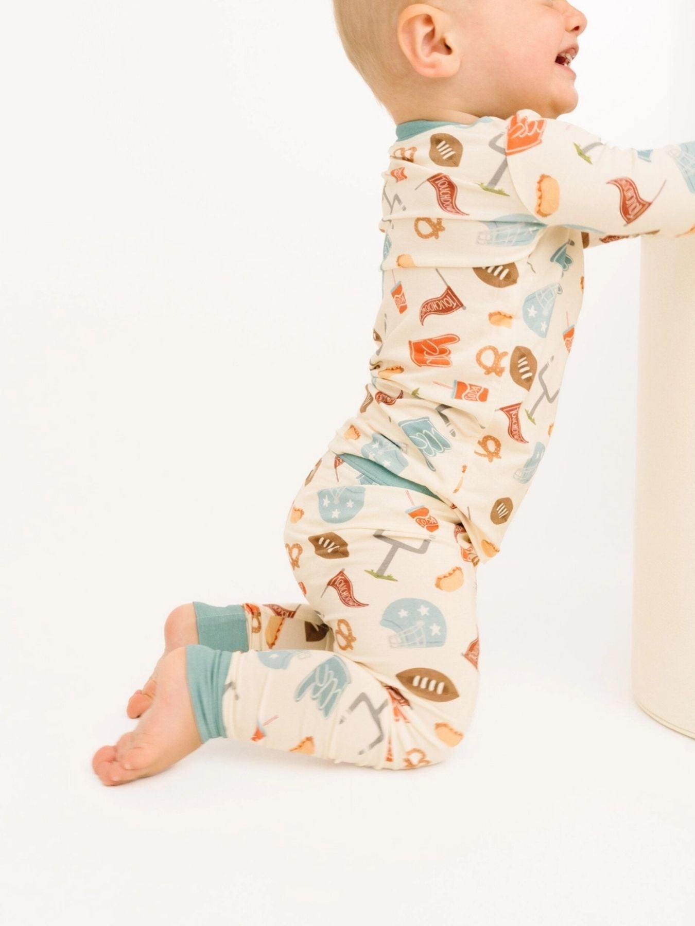 Toddler kneeling down wearing a  two-piece pajama set covered in playful football icons — helmets, pretzels, foam fingers, and more — paired with dusty blue trim at the collar and cuffs on a white background 