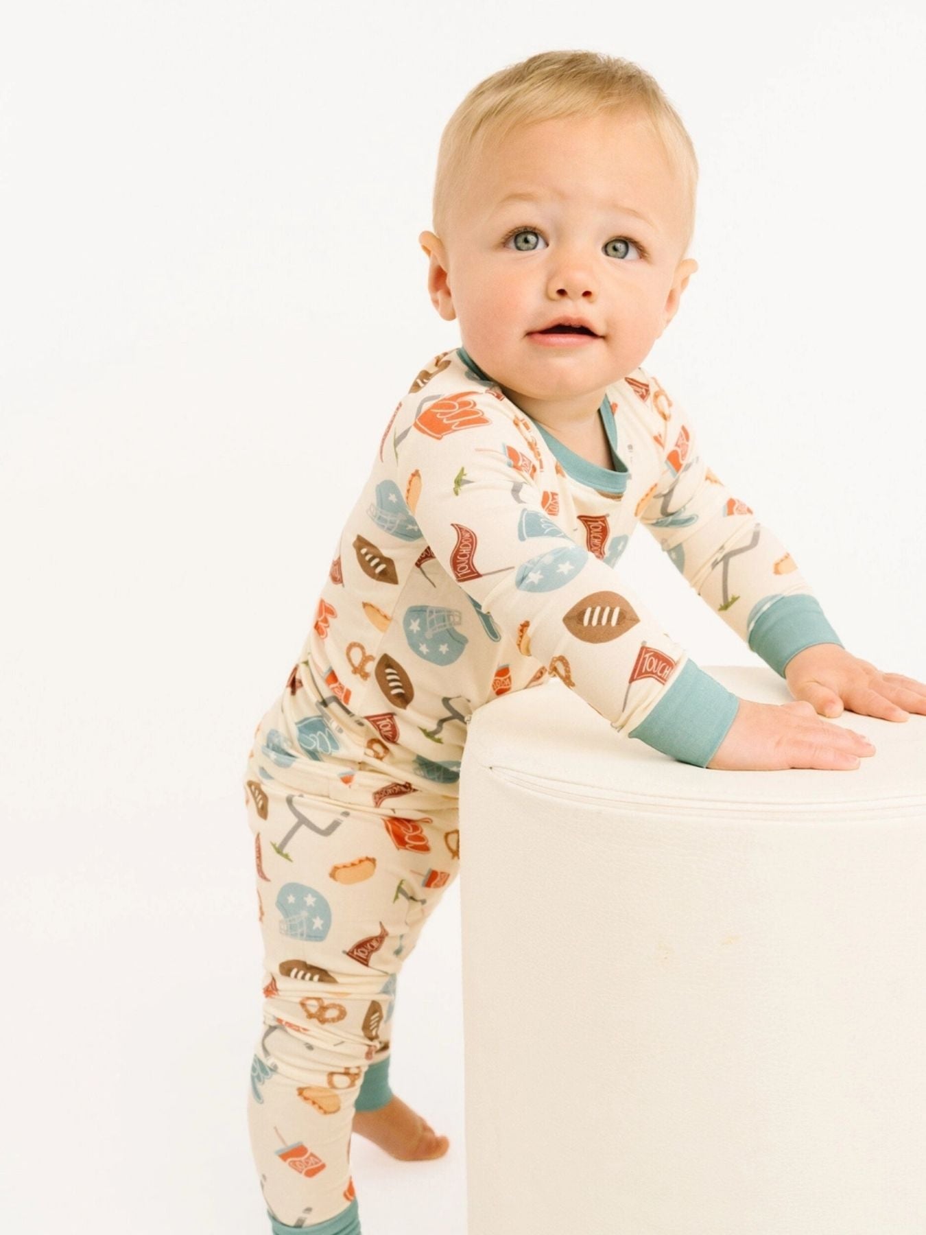 Toddler wearing a  two-piece pajama set covered in playful football icons — helmets, pretzels, foam fingers, and more — paired with dusty blue trim at the collar and cuffs on a white background 