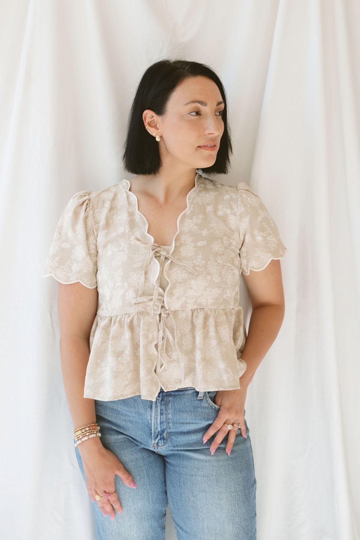 Model wearing the Floral Embroidered Front Tie Scalloped Peplum Top. Sweet and classy, this top can be dressed up easily or kept casual depending what you pair with it. 
