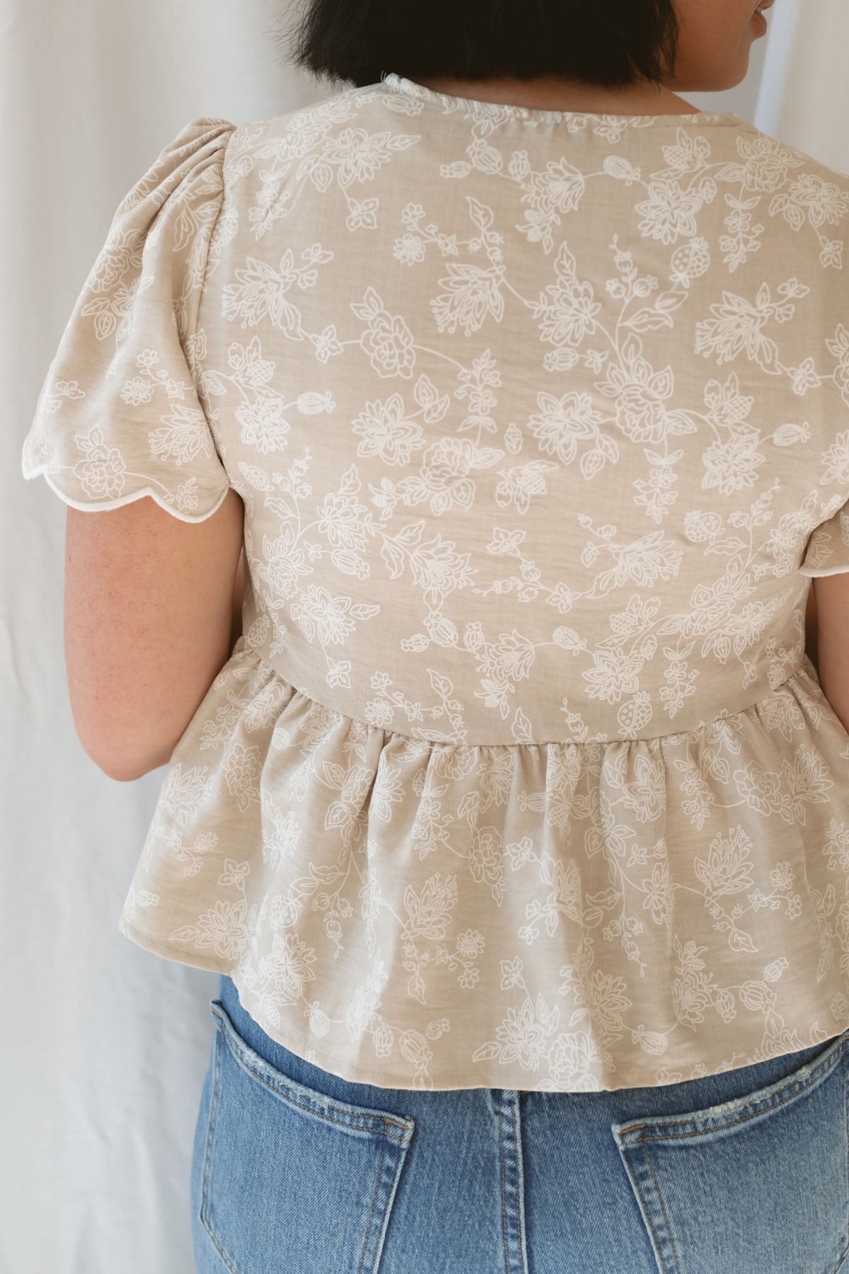 Back view of model wearing the Floral Embroidered Front Tie Scalloped Peplum Top. Sweet and classy, this top can be dressed up easily or kept casual depending what you pair with it. 