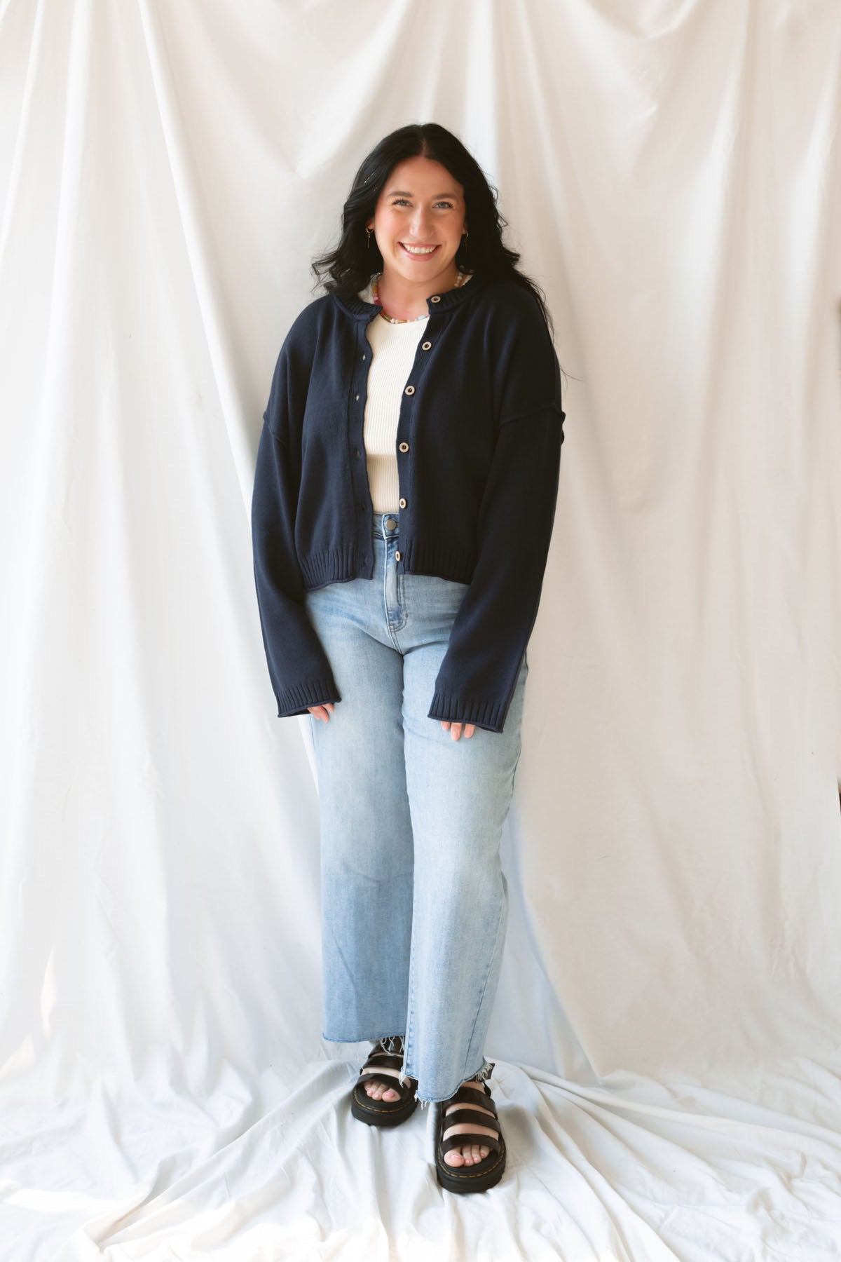 Model wearing the navy full button raw edge cardigan. Styled fully unbuttoned over a cream tank and paired with basic denim jeans. 
