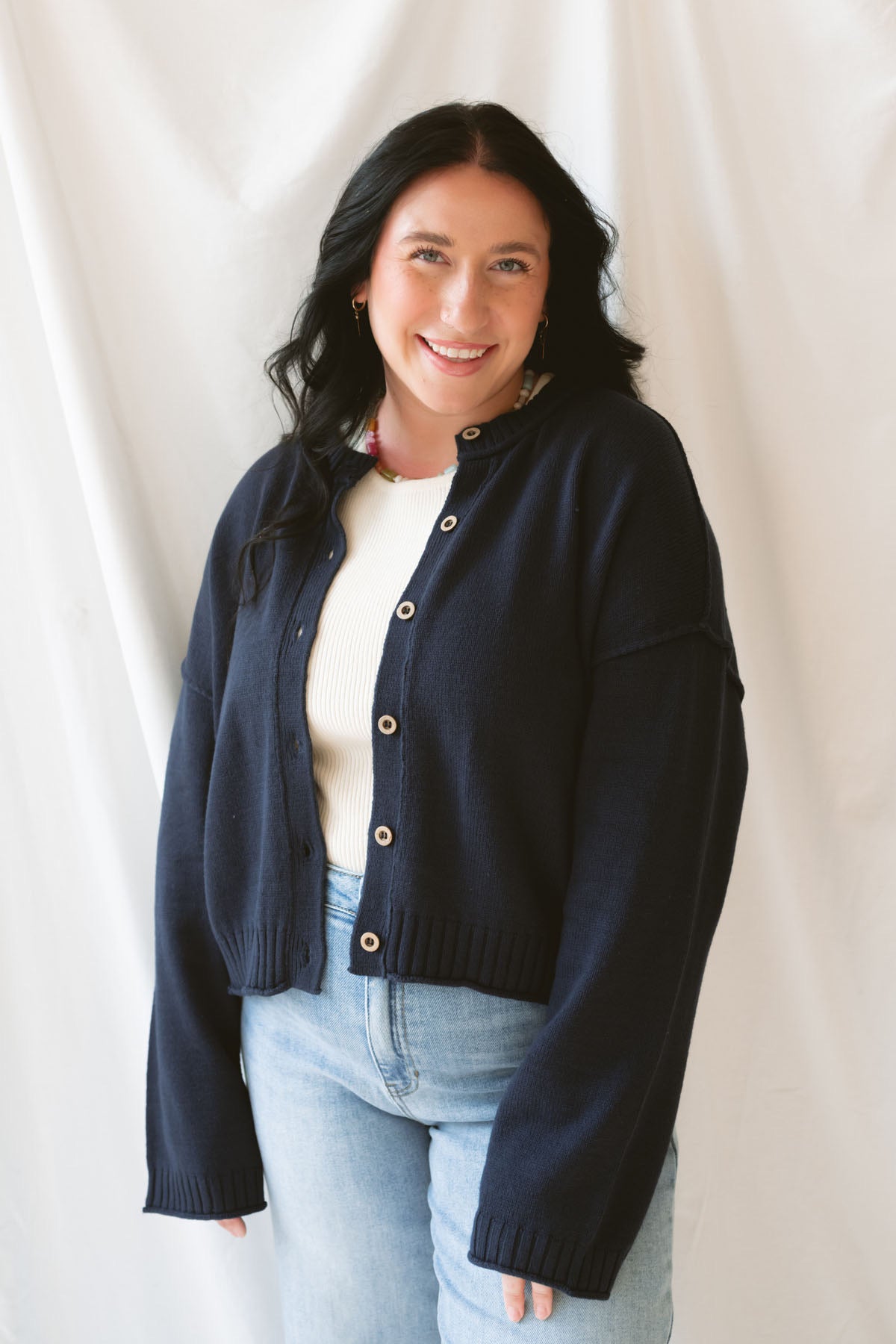 Model wearing the navy full button raw edge cardigan. Styled fully unbuttoned over a cream tank and paired with basic denim jeans. 
