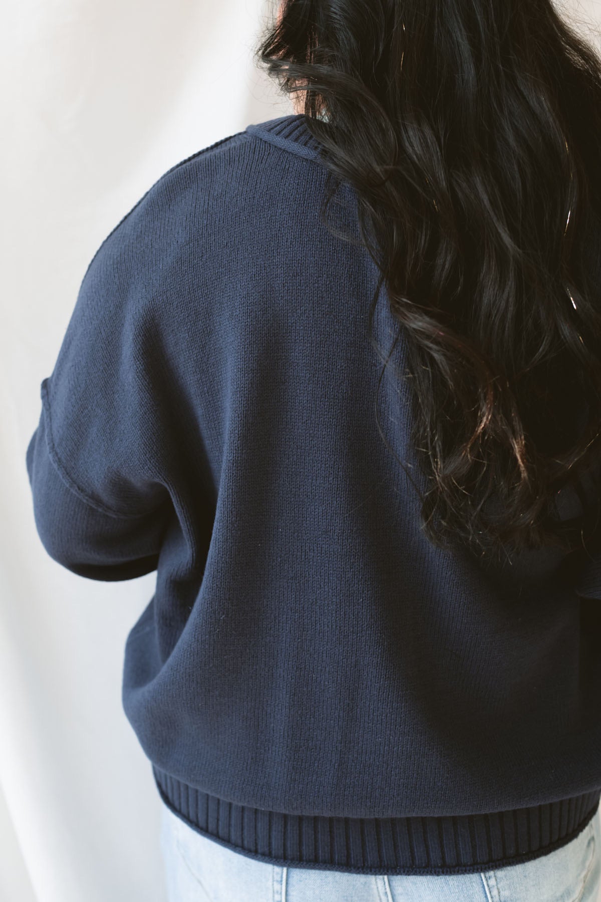 Back view of model wearing the navy full button raw edge cardigan. Styled fully unbuttoned over a cream tank and paired with basic denim jeans. 