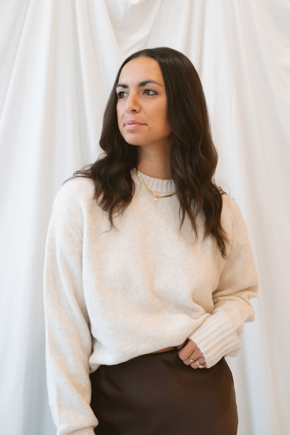 Model wearing a round neck solid sweater in light taupe. Styled with a brown satin skirt with lace trim.
