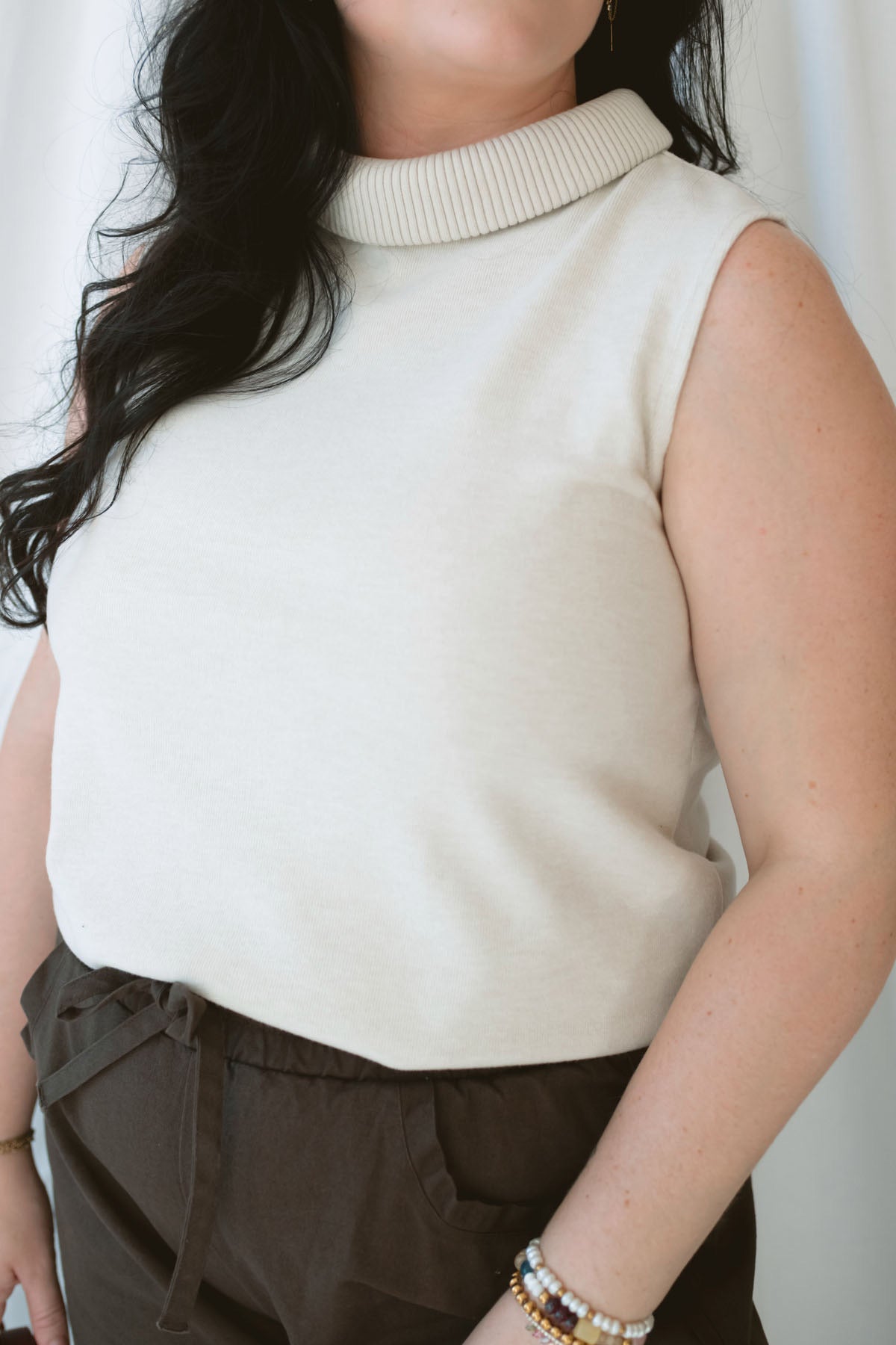 Model wearing sleeveless beige turtleneck. 