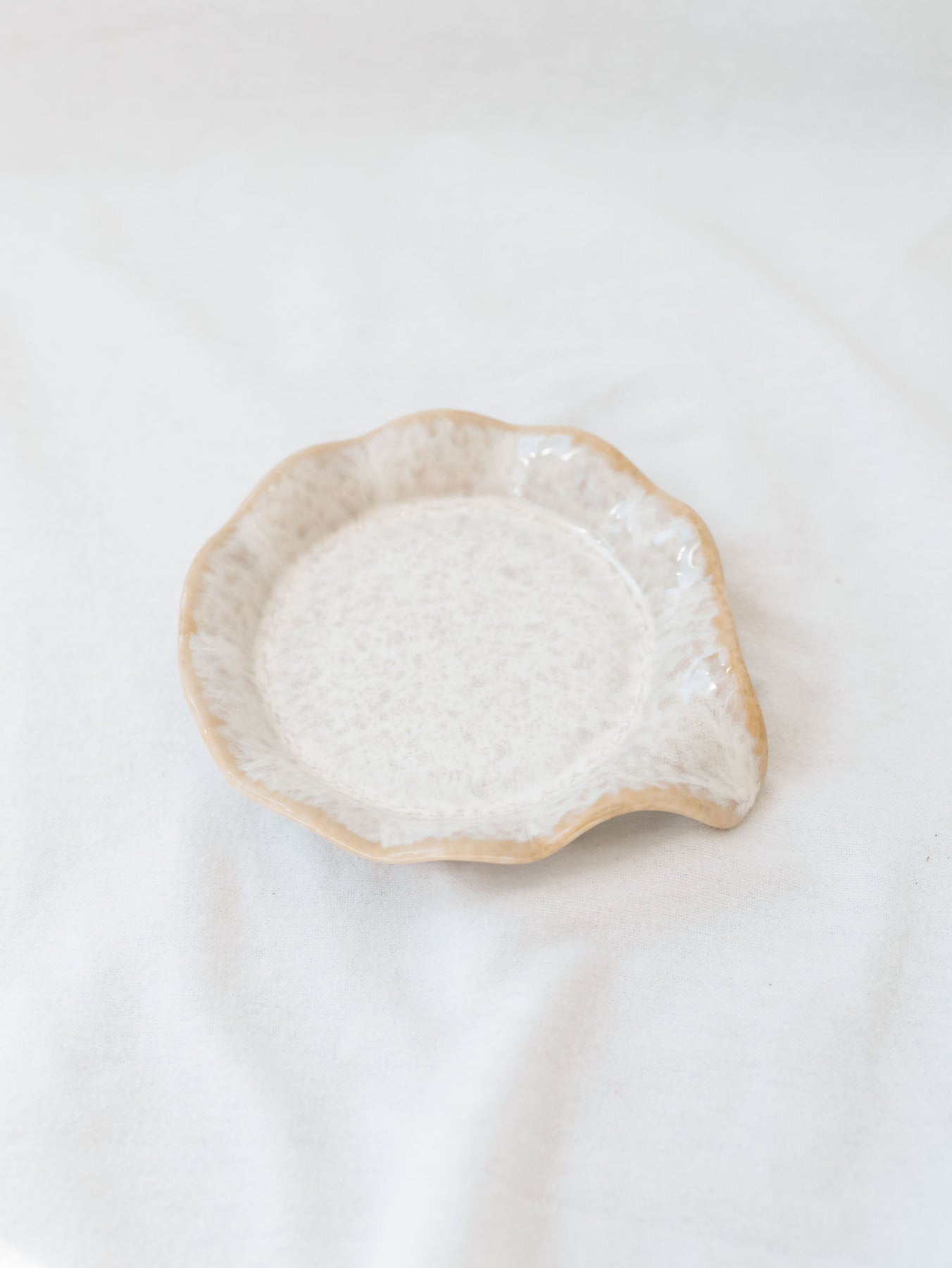 Beige scallop edge spoon rest on white linen backdrop. 