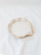 Beige scallop edge spoon rest on white linen backdrop. 