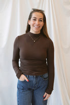 Chocolate brown long-sleeve knit turtleneck top for women featuring ruched waist detailing, a softly shaped silhouette, and an elevated everyday design.