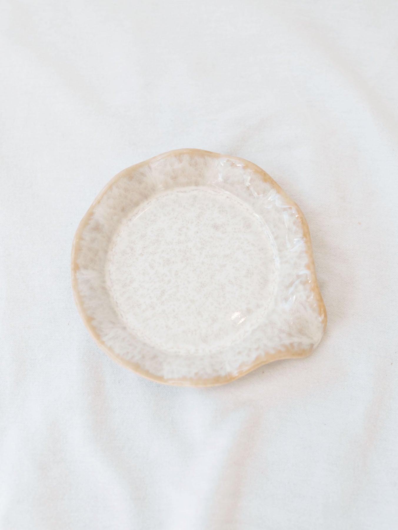 Beige scallop edge spoon rest on white linen backdrop. 