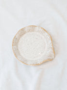 Beige scallop edge spoon rest on white linen backdrop. 