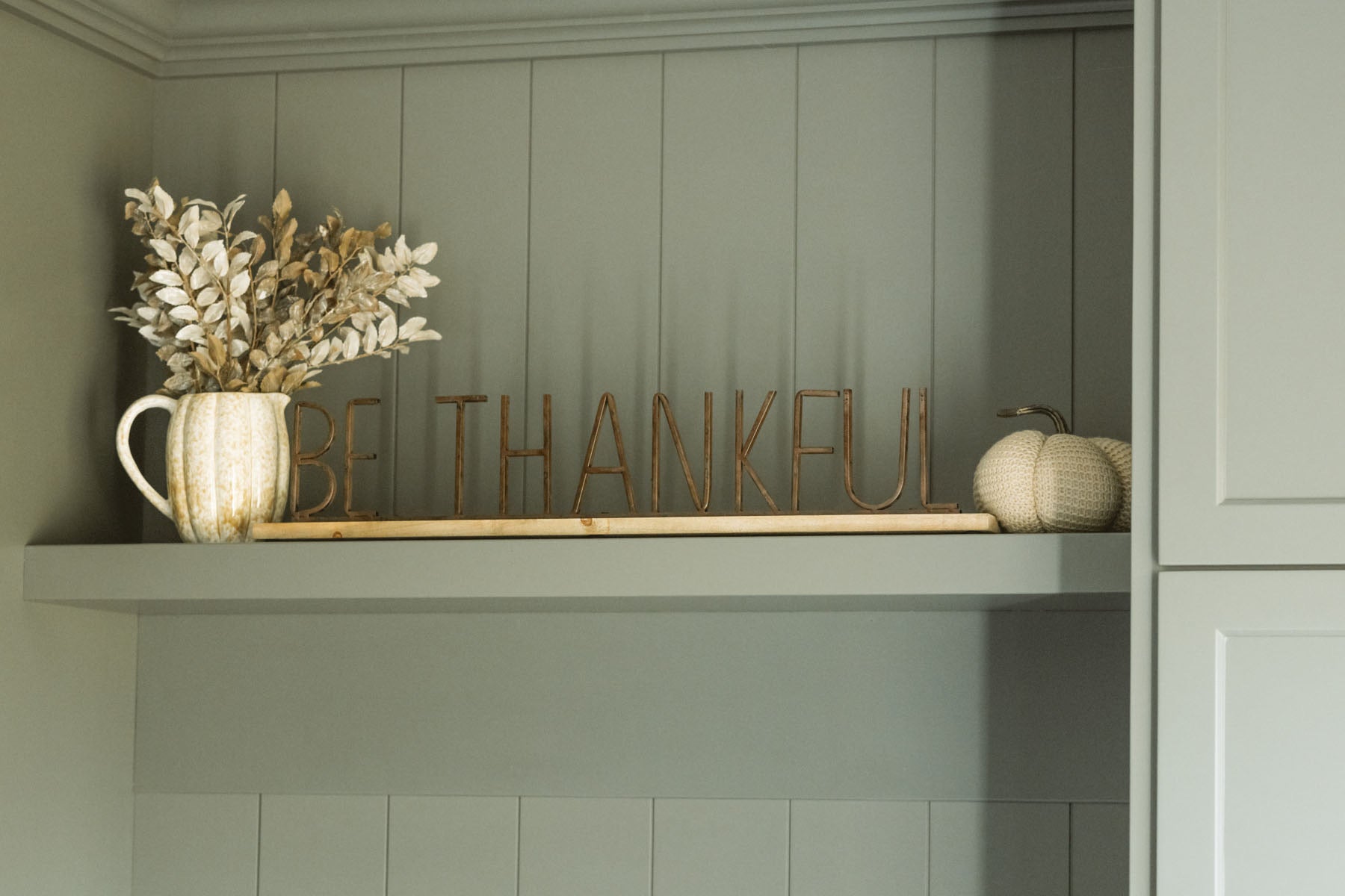 Be Thankful style metal and wood sign sitting on shelf styled with a pumpkin vase and foliage in addition to a knit beige pumpkin. 
