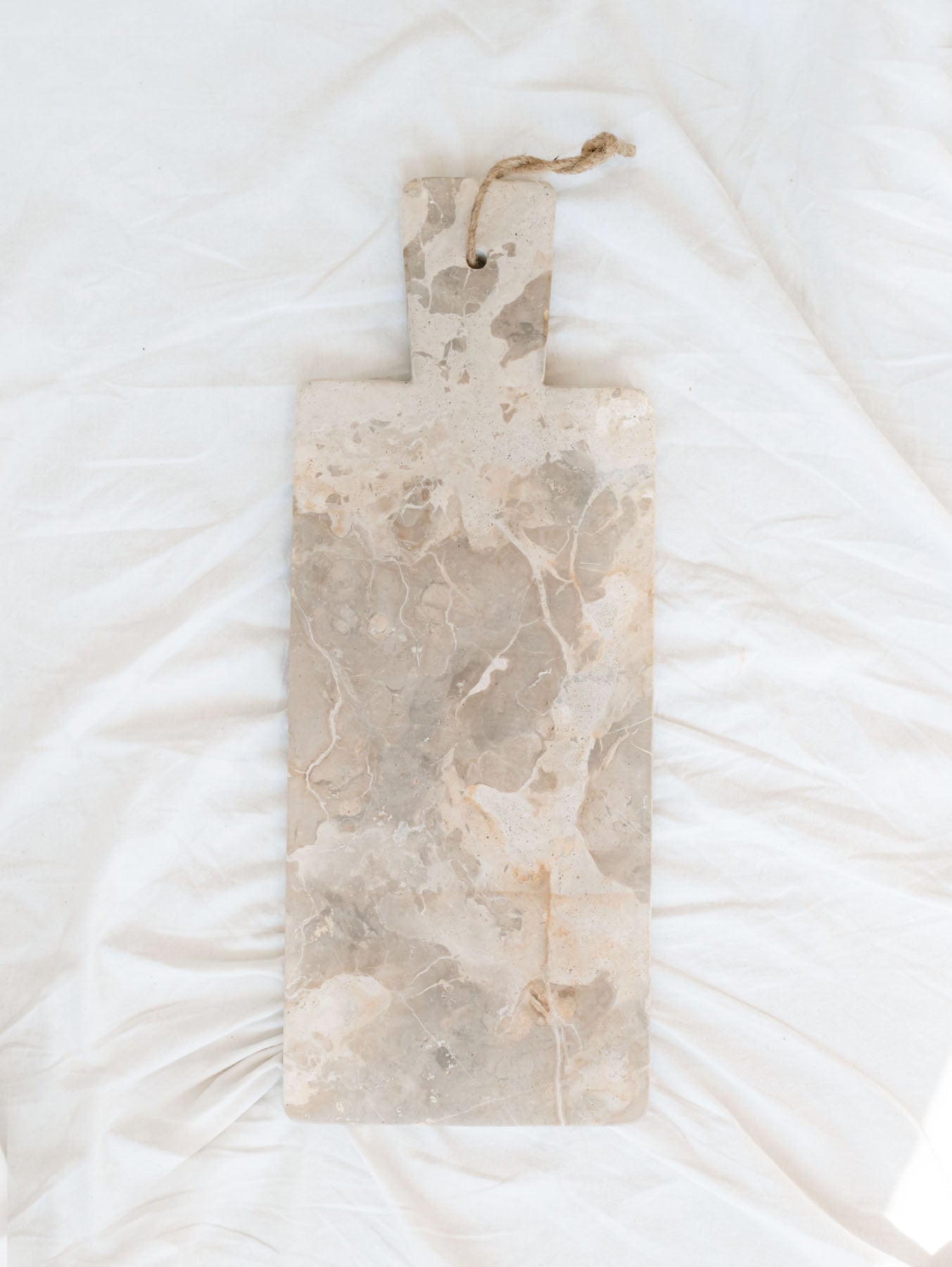 Beige marble rectangle cheese board with hanging rope laying on white linen backdrop. 